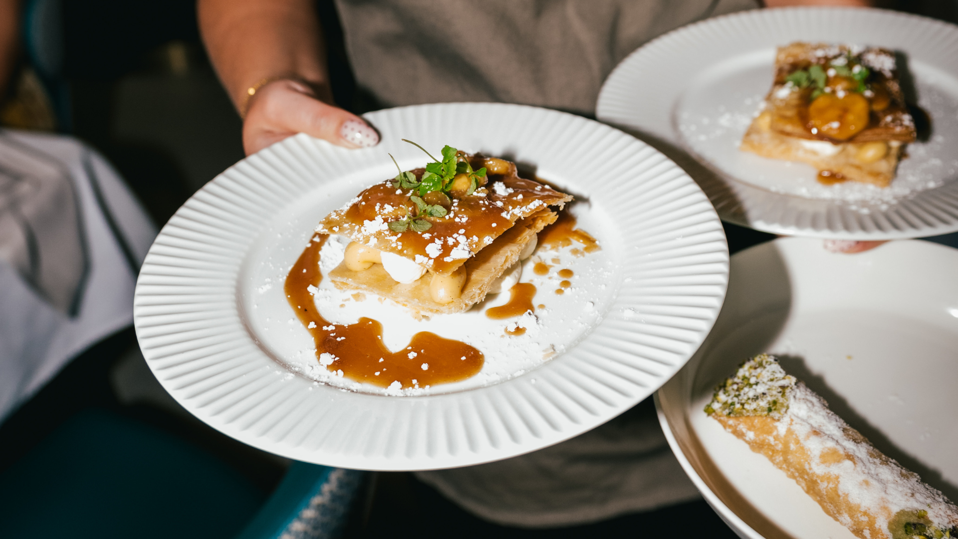 A person holding a white plate with a slice of dessert topped with caramel sauce, whipped cream, nuts, and herbs. There is another similar plate in the background and a third plate with a rolled pastry dusted with powdered sugar on a separate dish.