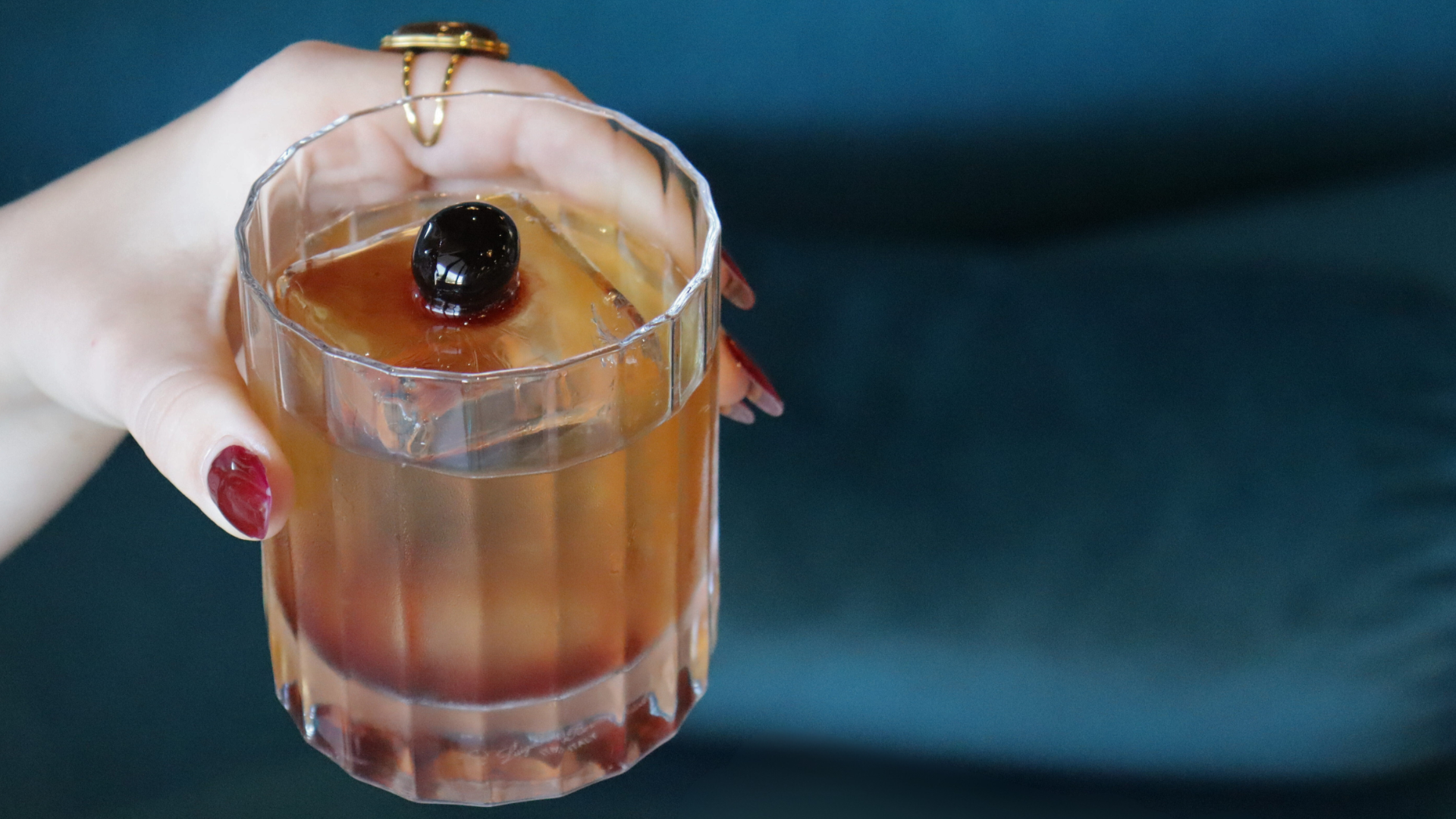 A hand with red nail polish holding a glass of iced coffee with a black cherry on top, against a navy blue background.