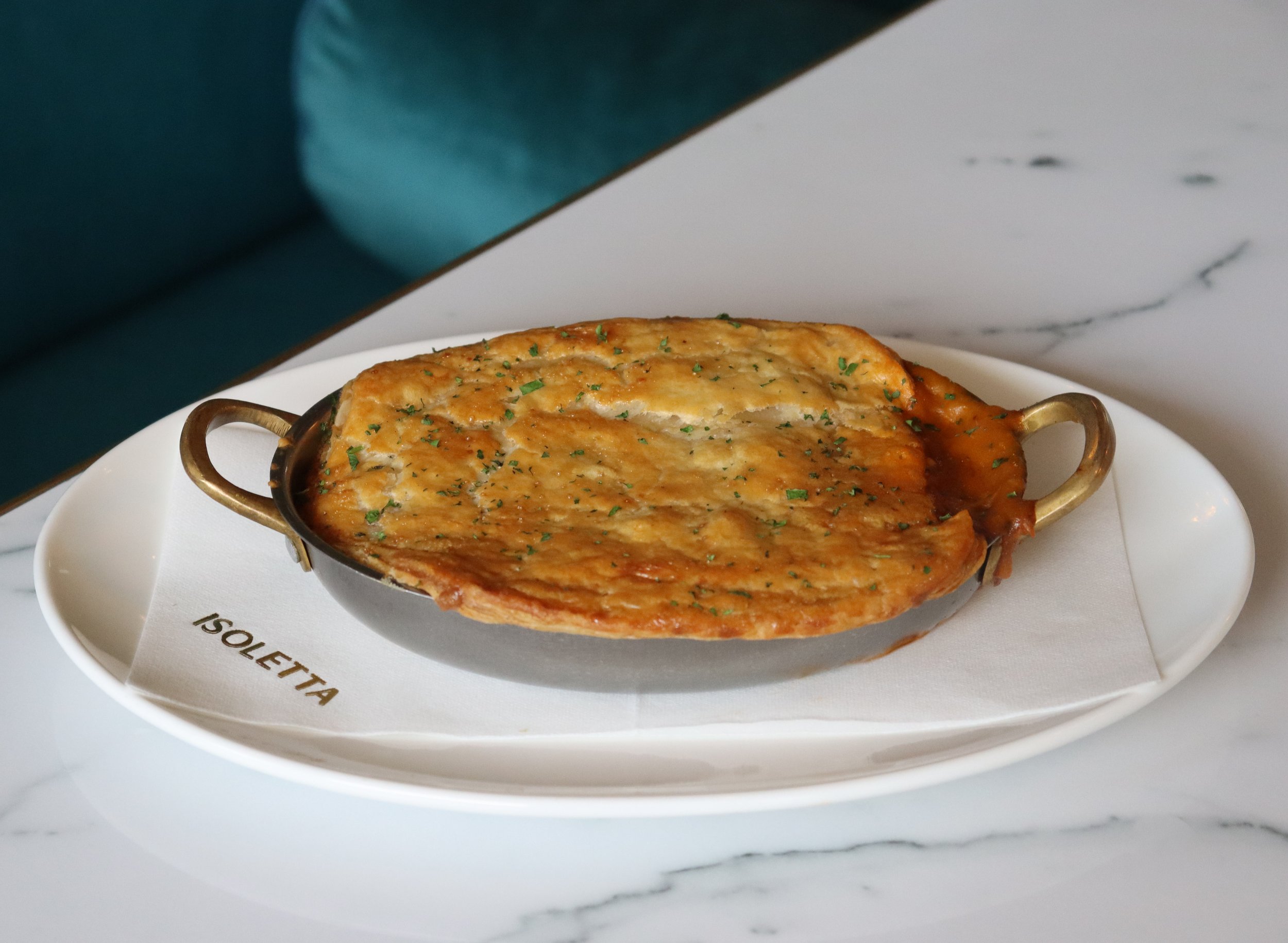 A silver dish filled with baked fish, garnished with chopped herbs, resting on a white plate with gold handles, placed on a white marble surface.