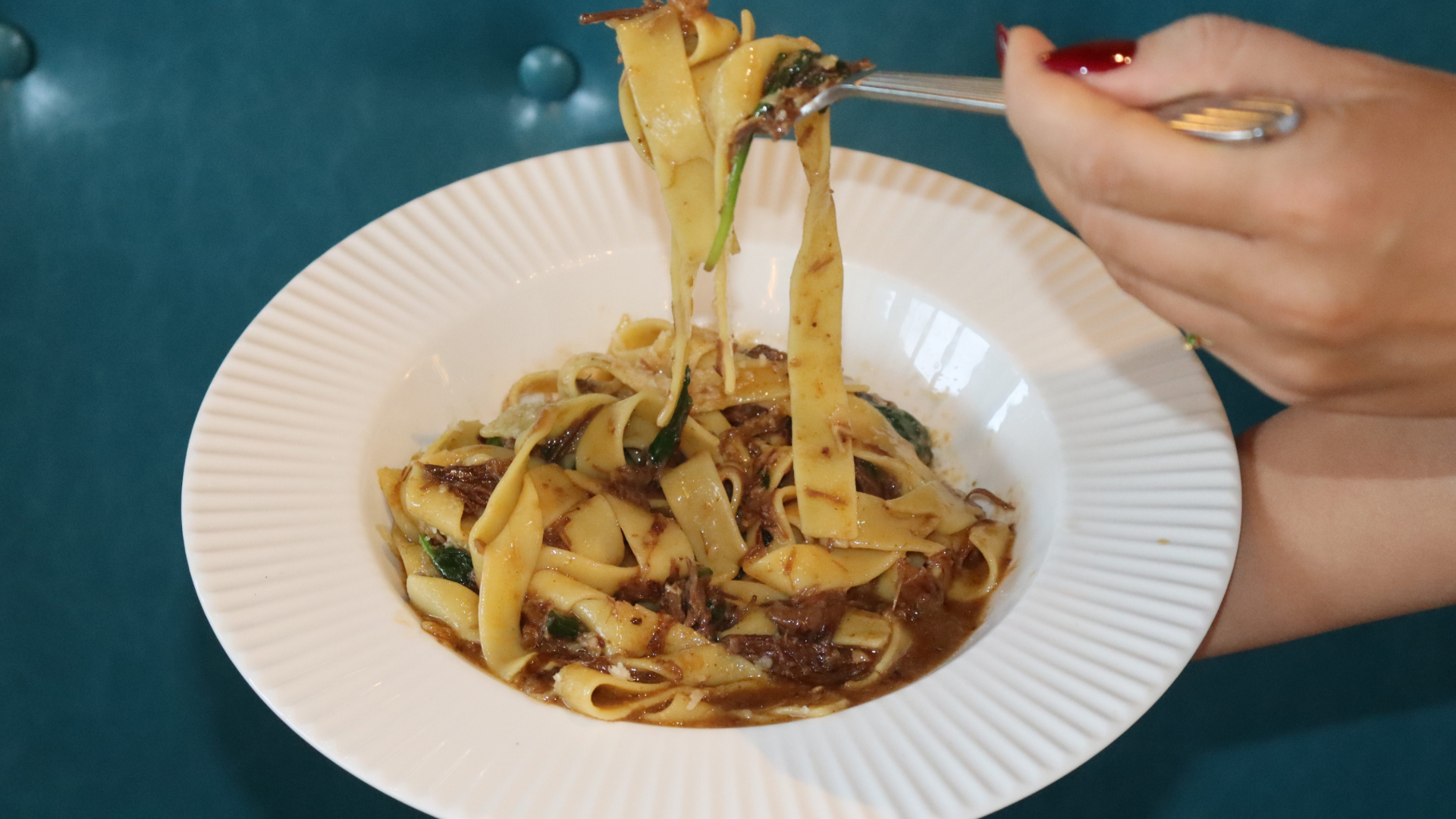 Person holding a fork of pasta over a white plate with pasta and sauce on a green table.