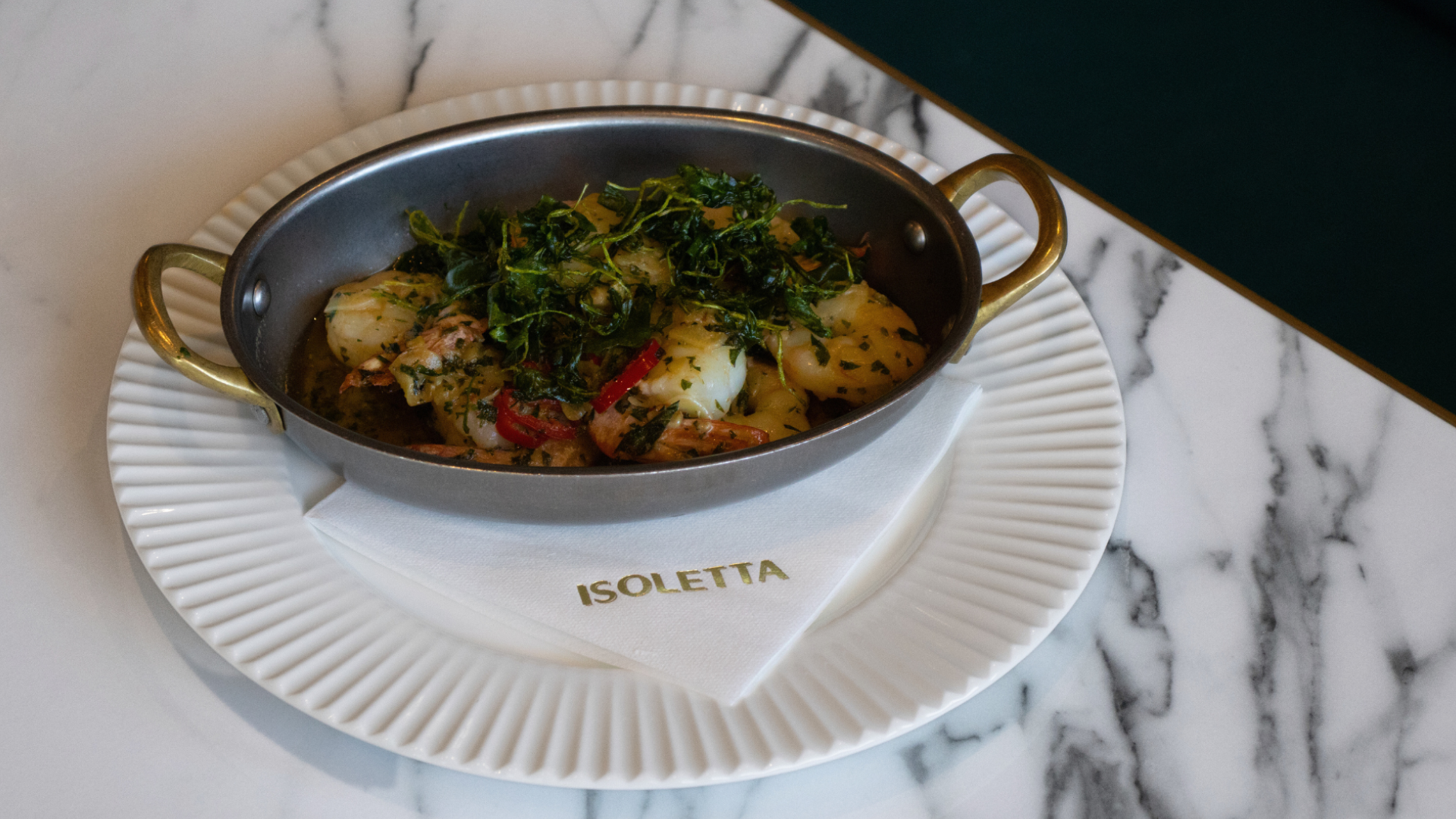 Dish of seafood with green herbs on a white plate with a napkin that says 'ISOLETTA', placed on a marble surface
