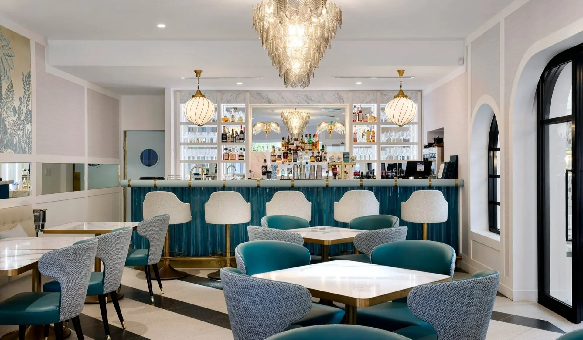 Interior of a stylish, well-lit bar and restaurant with a marble countertop, barstools, and decorative lighting fixtures, including chandeliers and pendant lights, along with a back bar stocked with various bottles and glassware.
