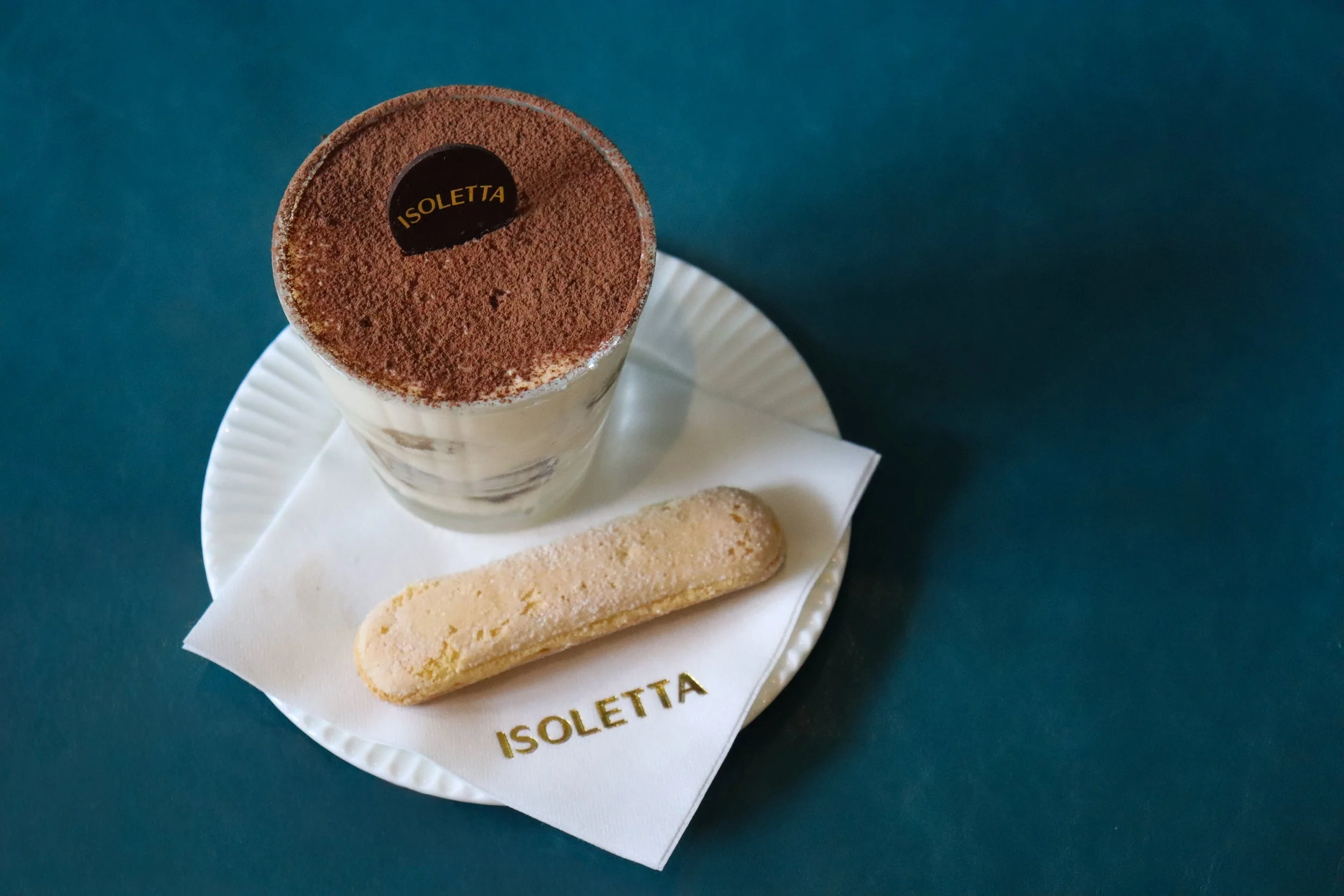 A dessert cup topped with cocoa powder and a chocolate medallion labeled 'ISOLETTA.' On a white plate, there is a ladyfinger cookie and a piece of sponge cake, with a napkin underneath.