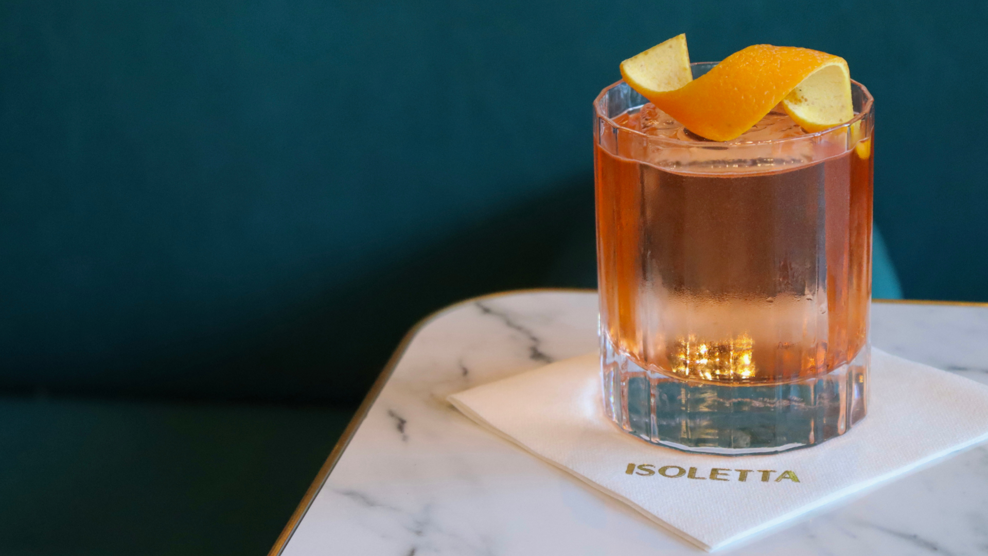A glass of pink cocktail with orange peel garnish on a white napkin with gold lettering that reads 'ISOLETTA' on a marble table