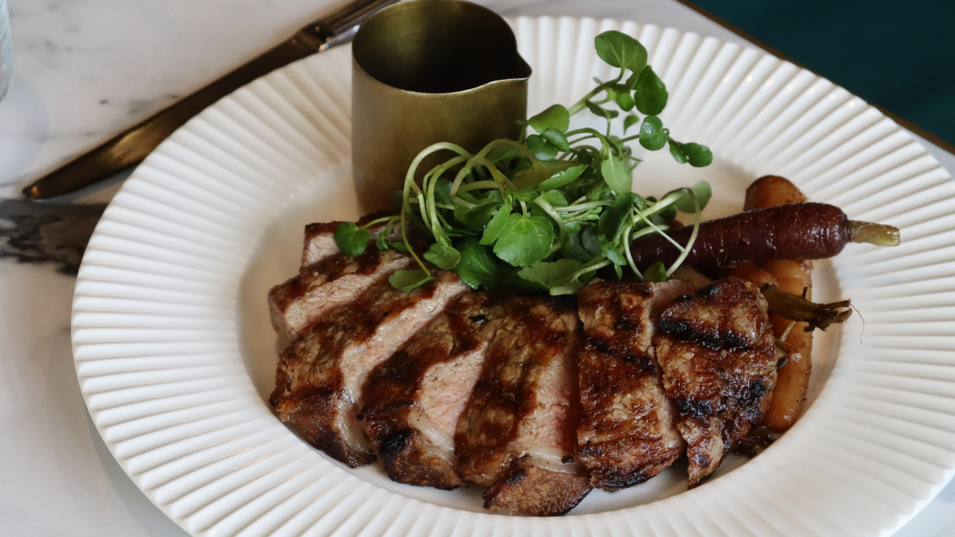 A white plate with grilled meat, small carrots, and microgreens, with a small container of sauce or dressing on the side.