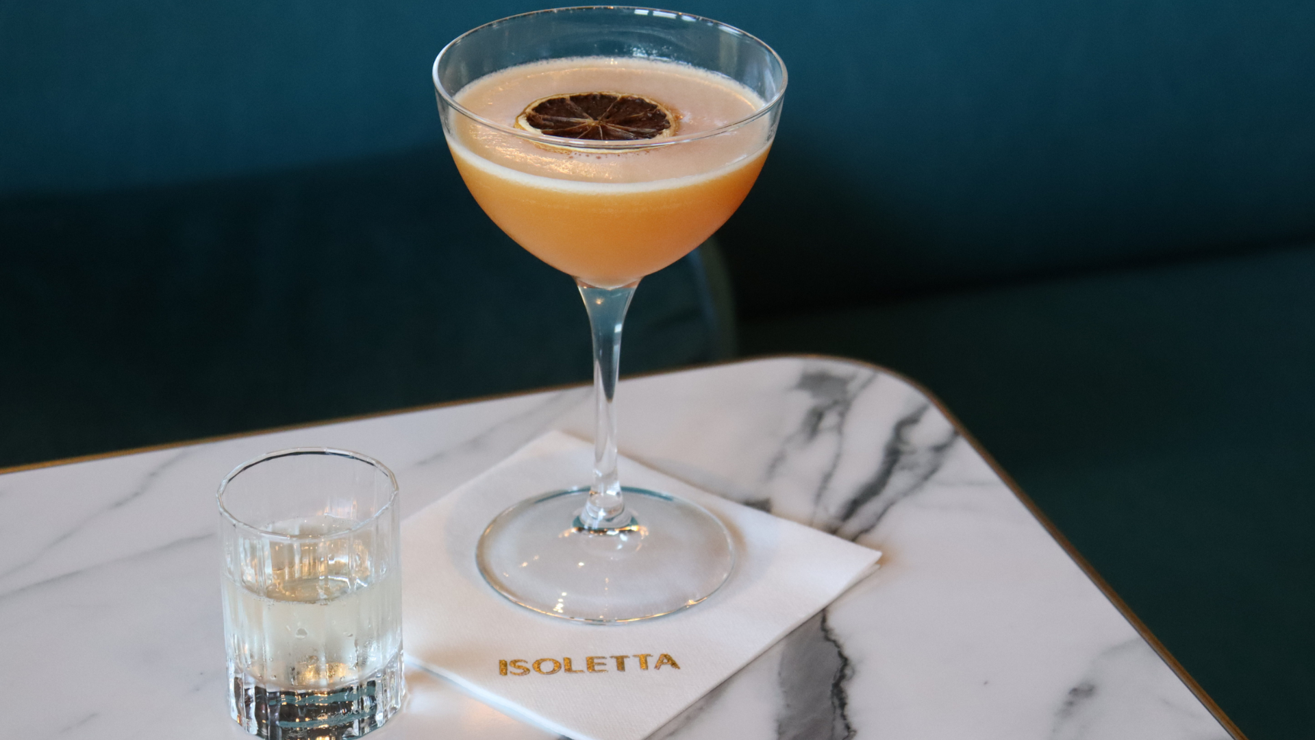 A pink cocktail with a dried lemon slice garnish in a martini glass, alongside a shot glass of clear liquid on a marble table with a white 'ISOLETTA' napkin.