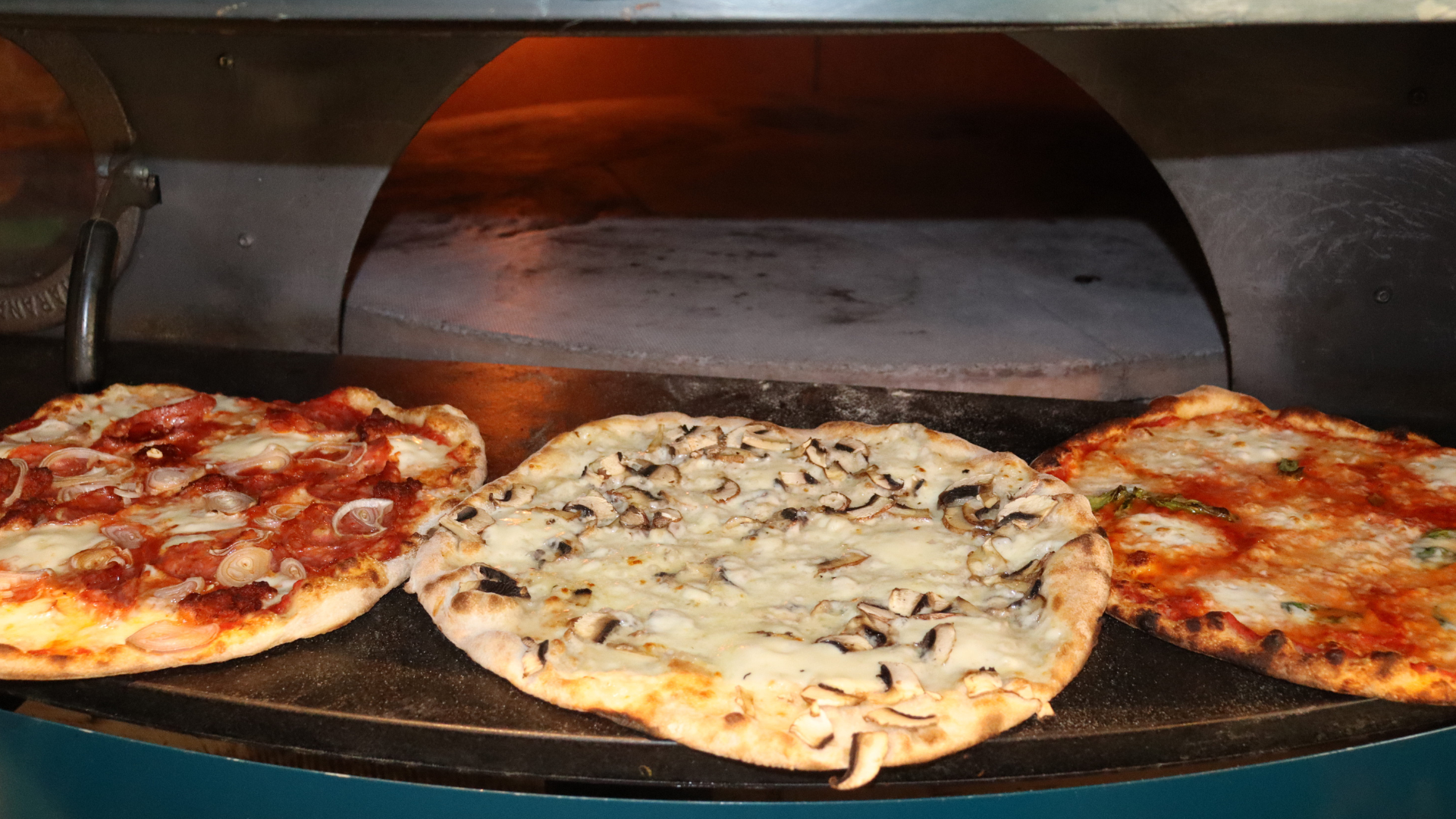 Three pizzas cooking in a pizza oven, with one pizza topped with onions and tomato, the second with mushrooms and cheese, and the third with cheese and green herbs.