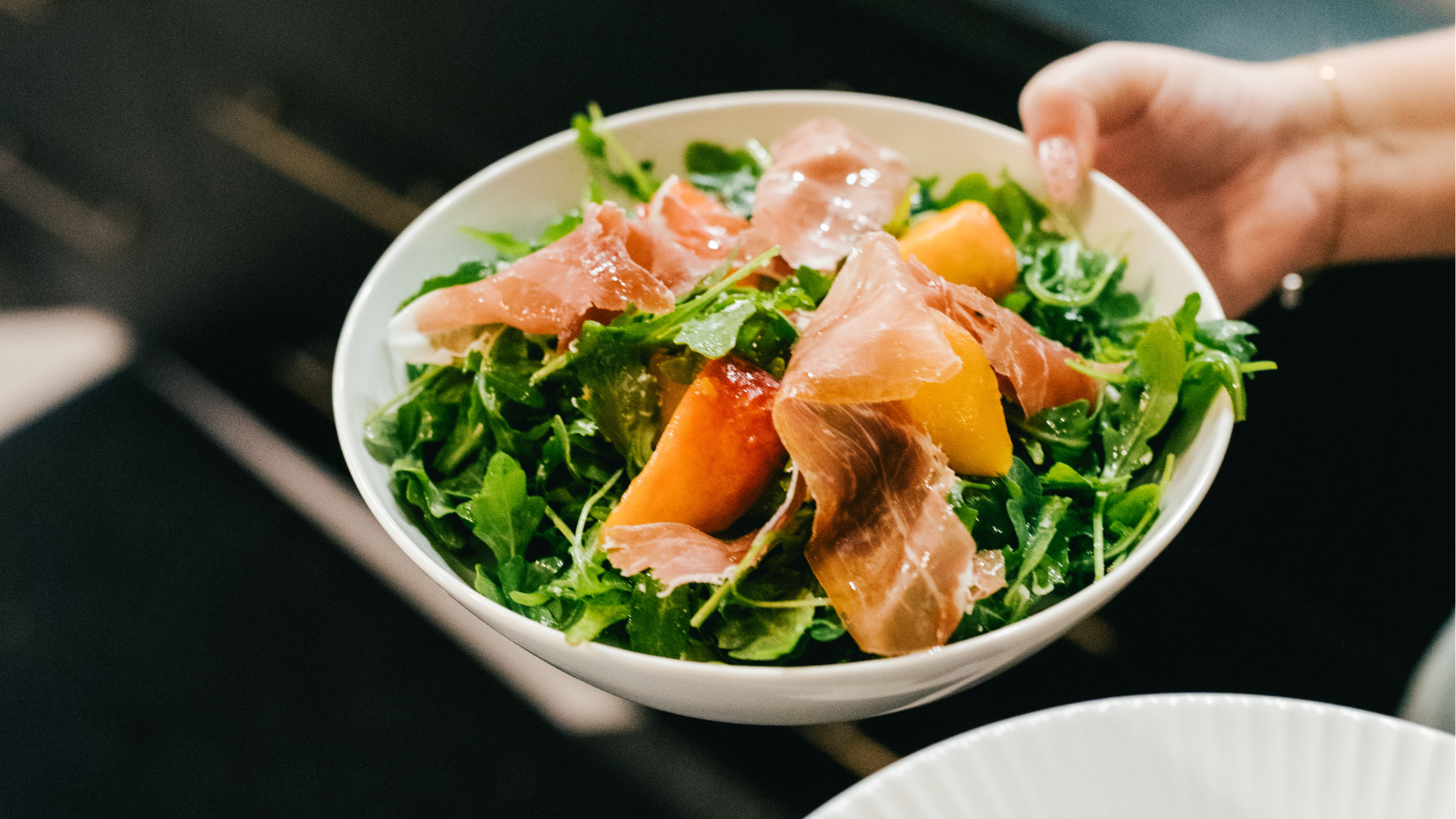 A white bowl filled with mixed greens, slices of prosciutto, and yellow peaches, held by a person's hand.