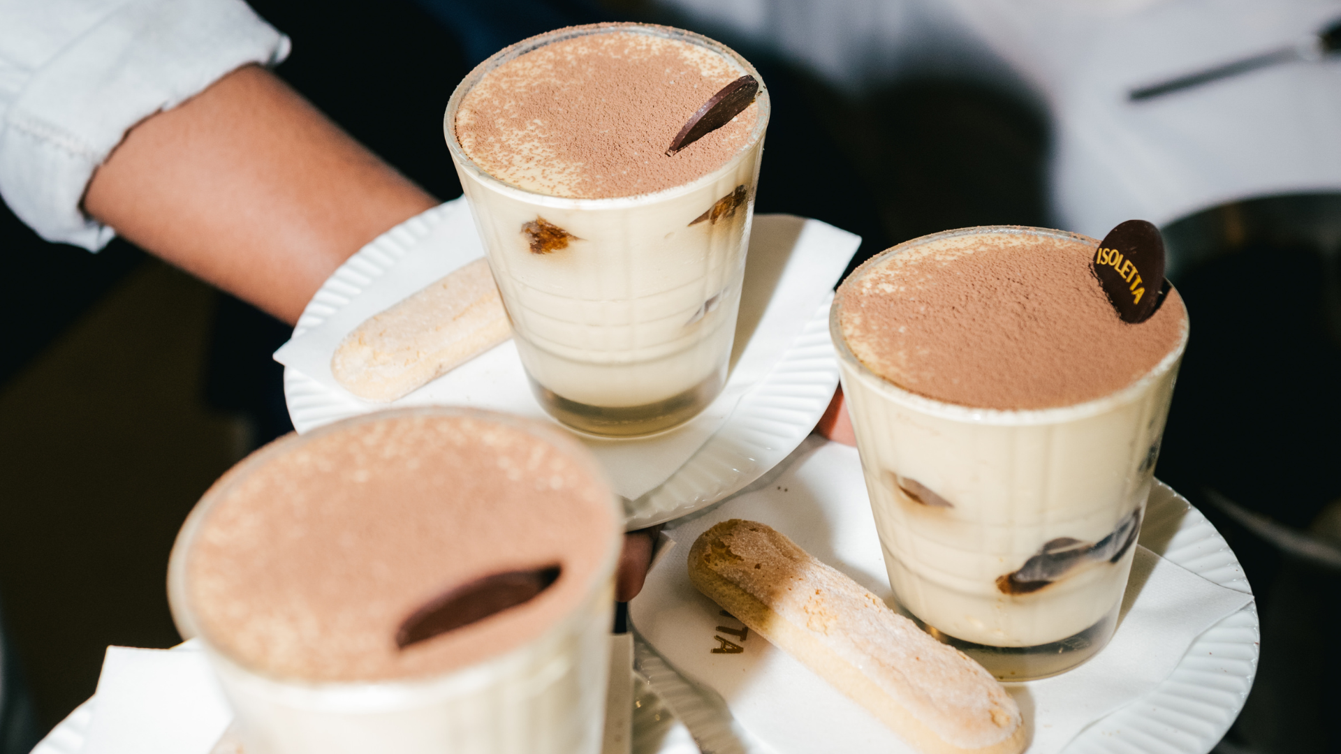 Three glasses of tiramisu dessert with cocoa powder and chocolate decoration, served on white plates with ladyfinger biscuits.