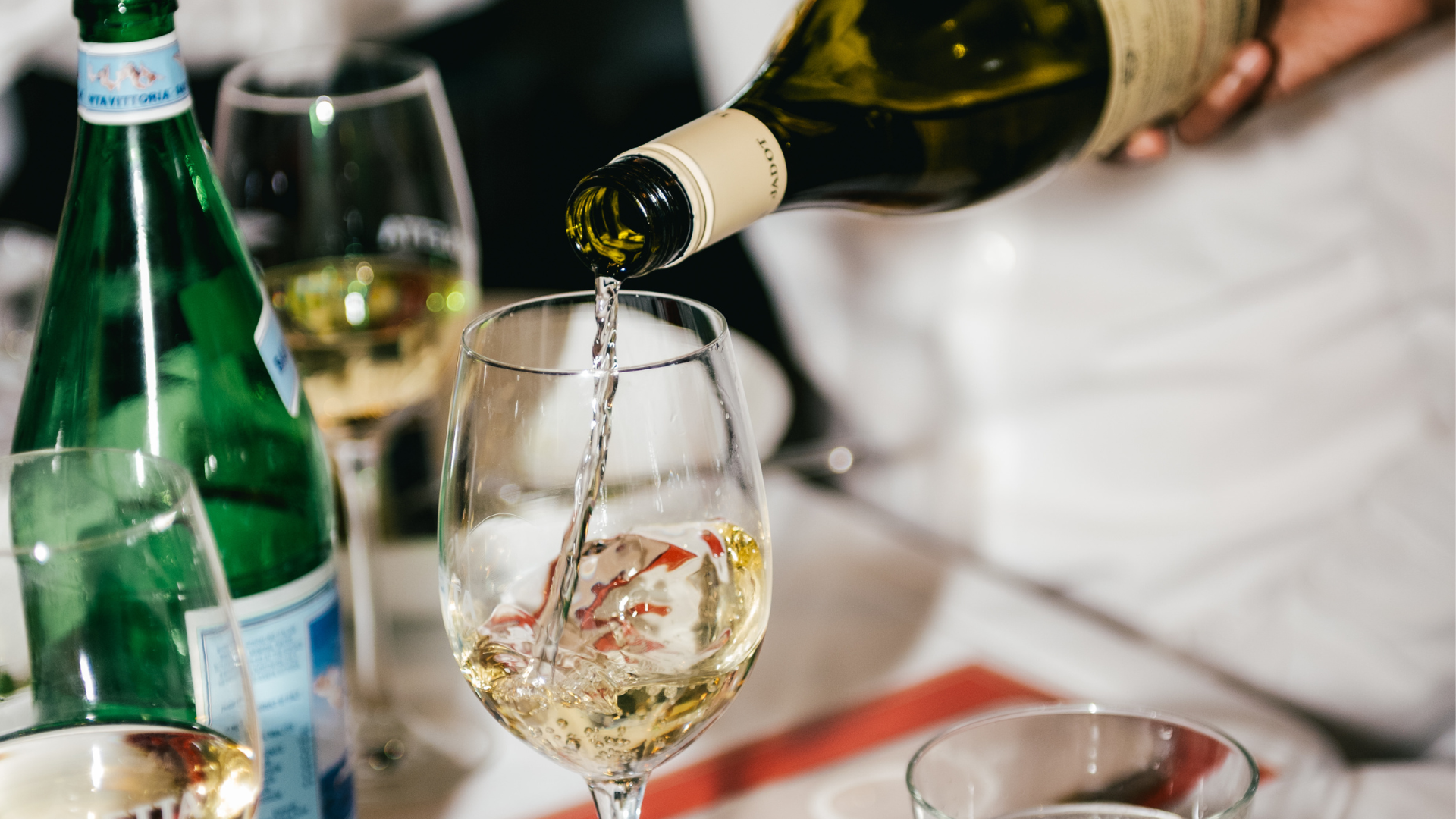 A person pouring white wine into a glass at a table with other glasses and a water bottle.