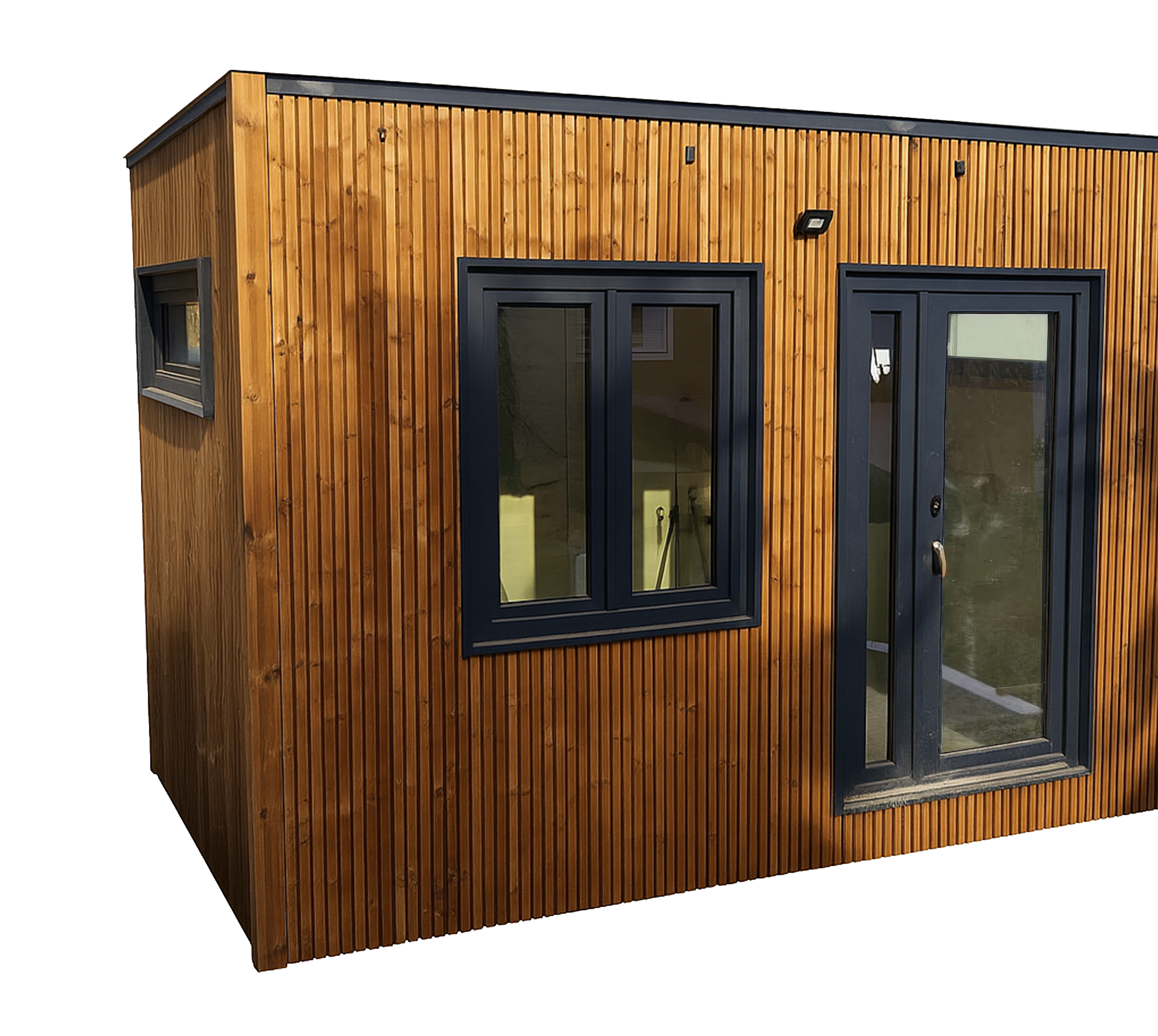 A small modern building with brown wooden exterior siding, black window frames, a glass door, and a small window on the side, all set against a dark background.