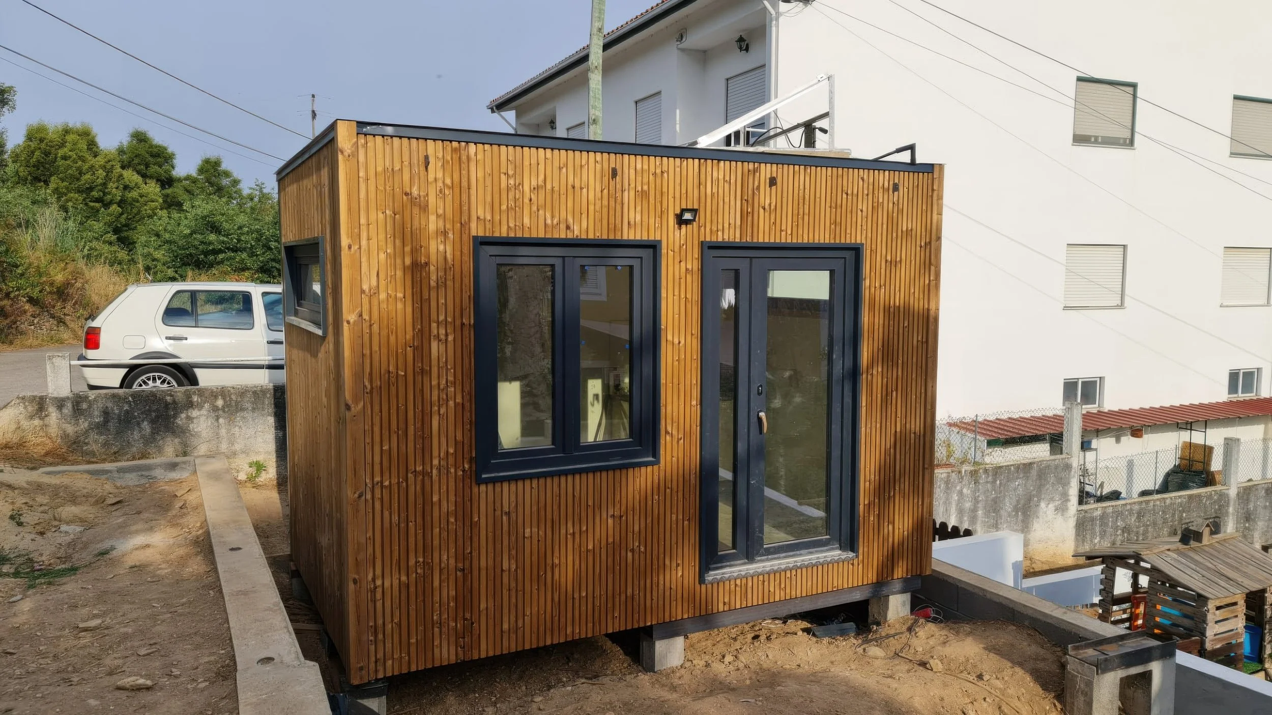 A small modern wooden building with black framed windows and door, built on concrete blocks, situated outdoors near a white building and parked car.