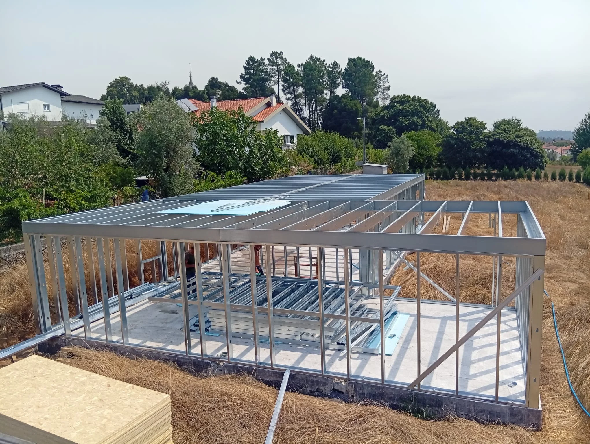 Construction site with a steel frame structure of a building in progress, surrounded by dry grass and several trees and houses in the background.