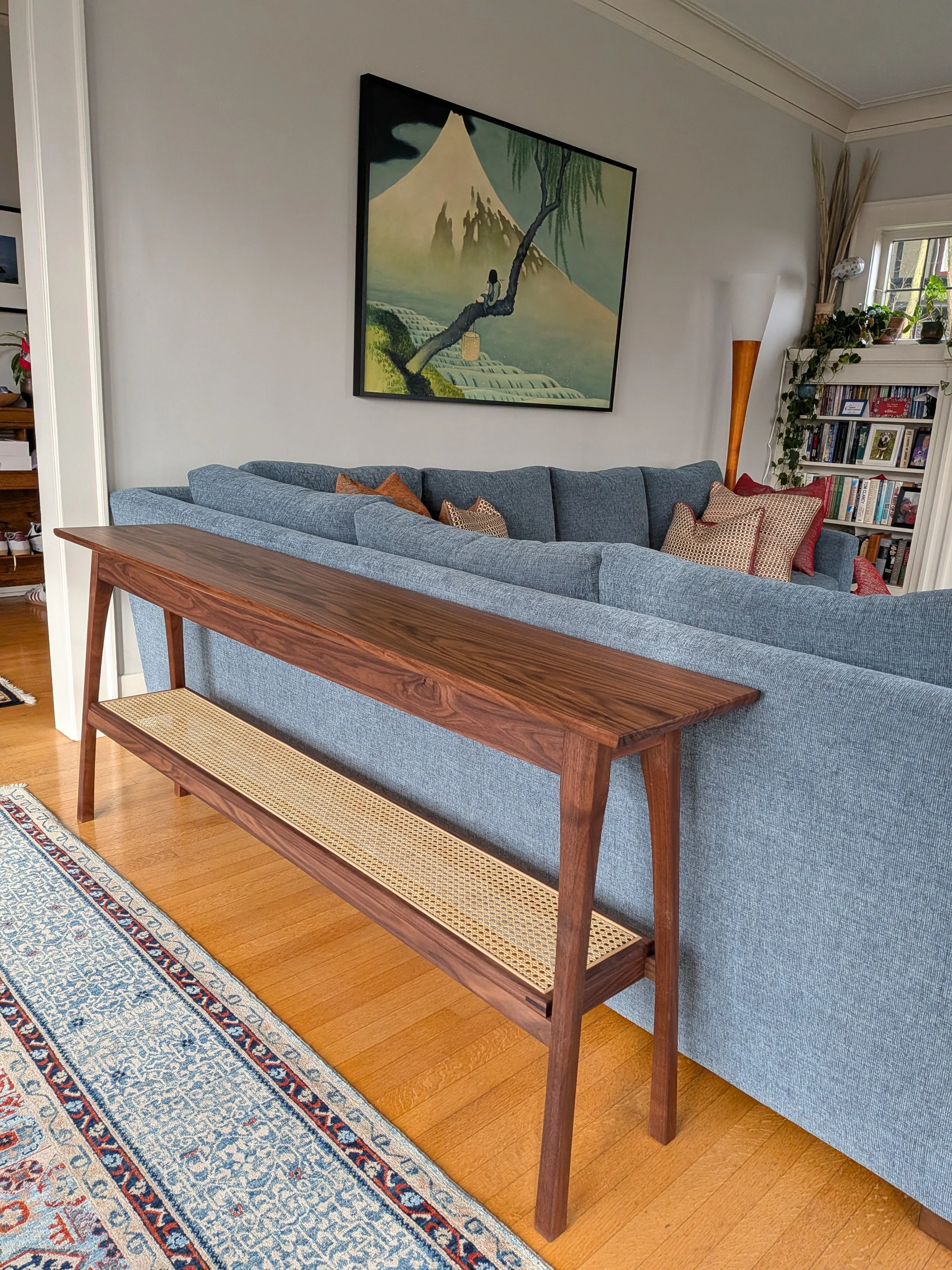 Custom walnut console table with splayed legs and cane shelf behind gray sectional