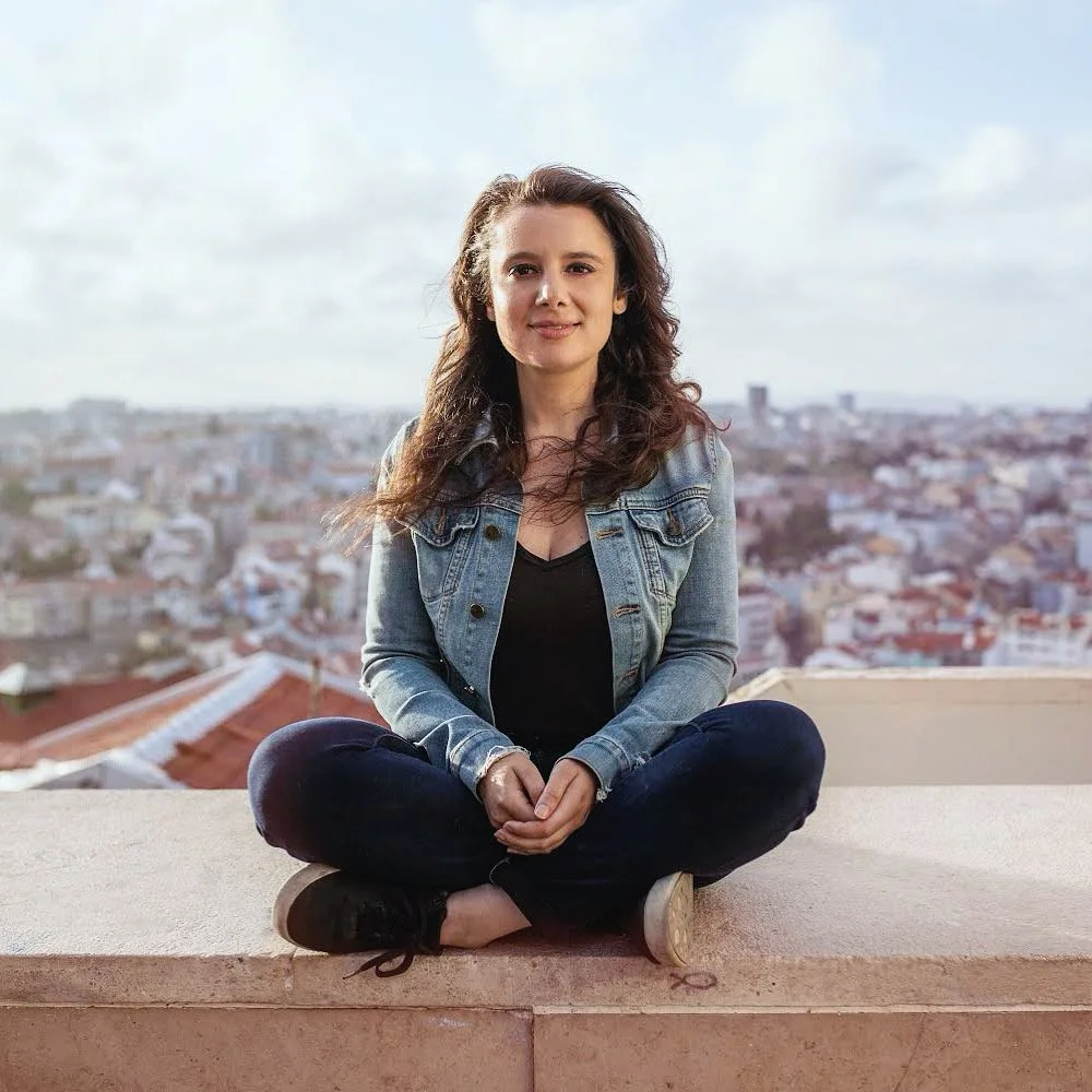A woman sitting cross-legged on a ledge with a cityscape in the background.