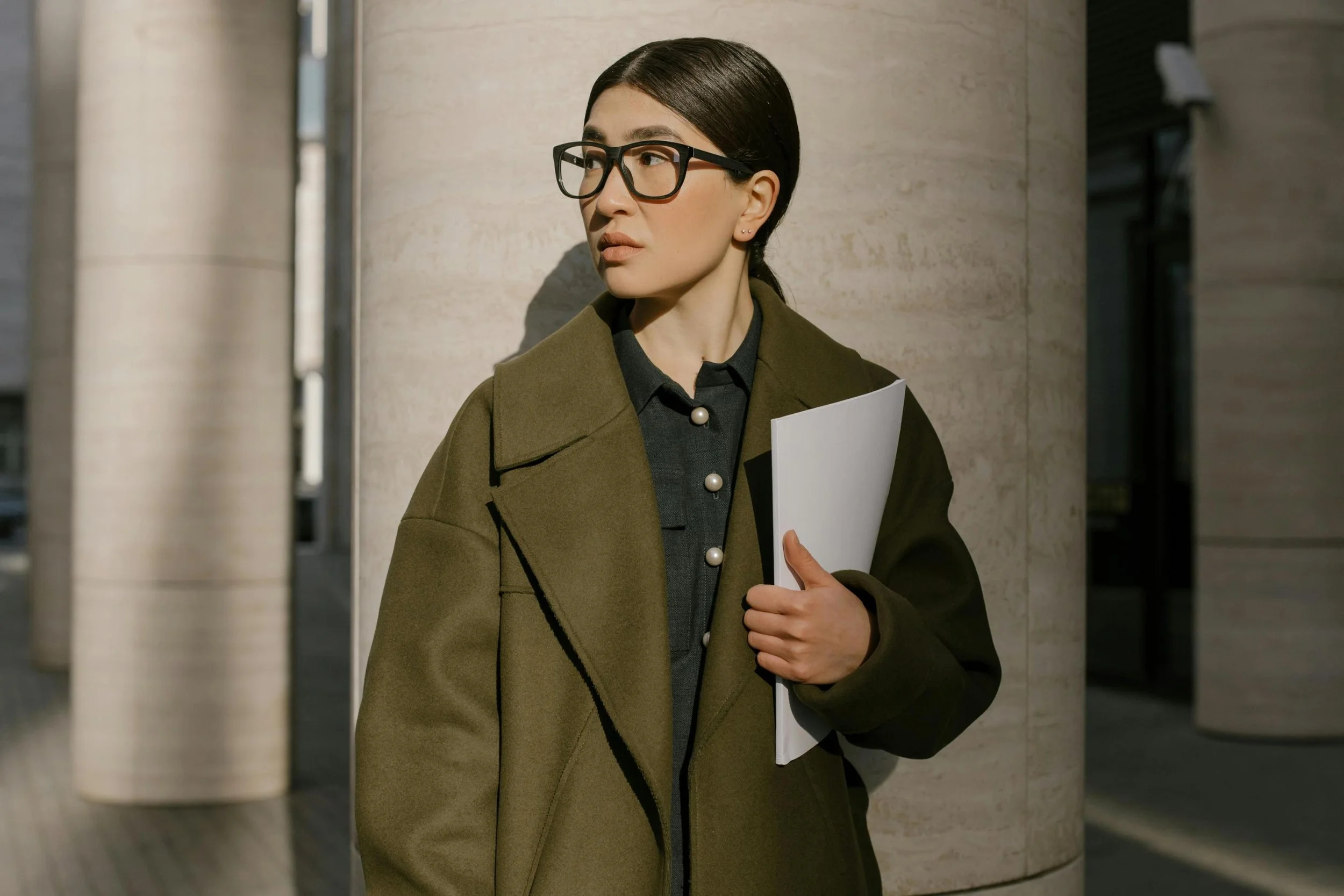 A woman with brown hair, wearing glasses, a dark coat, and holding a folder, leaning against a beige pillar outside in an urban setting.