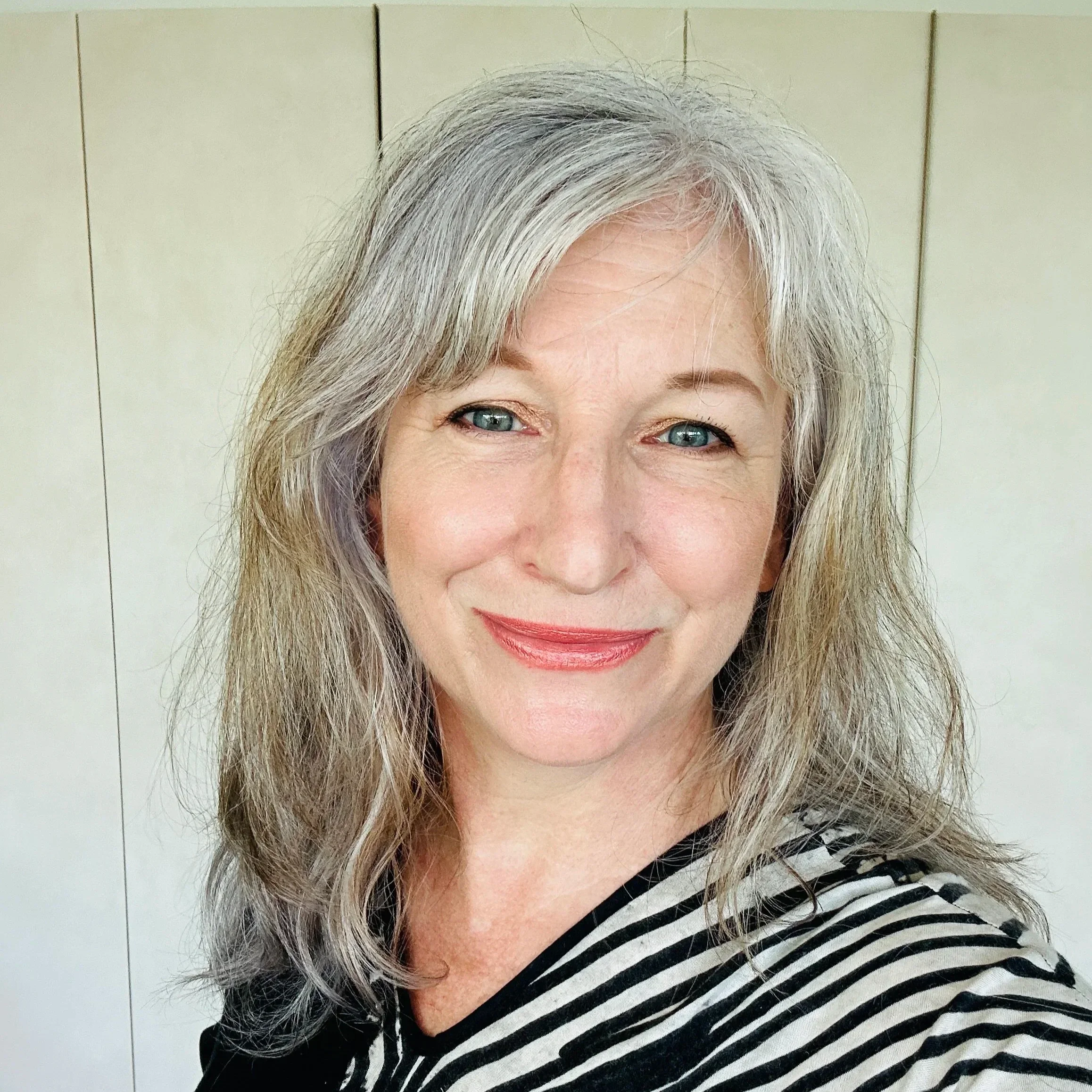 A warm, smiling artist Hilary Lee in her 50s with gray wavy hair and blue eyes, encouraging you to live outside the box