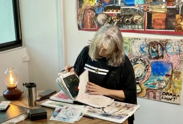 An artist in her 50s with white hair is sitting in a chaotic studio , looking at a sketch book and several sketches or notebooks. There is a window on the left, a lamp, and colorful abstract artwork on the wall behind her.