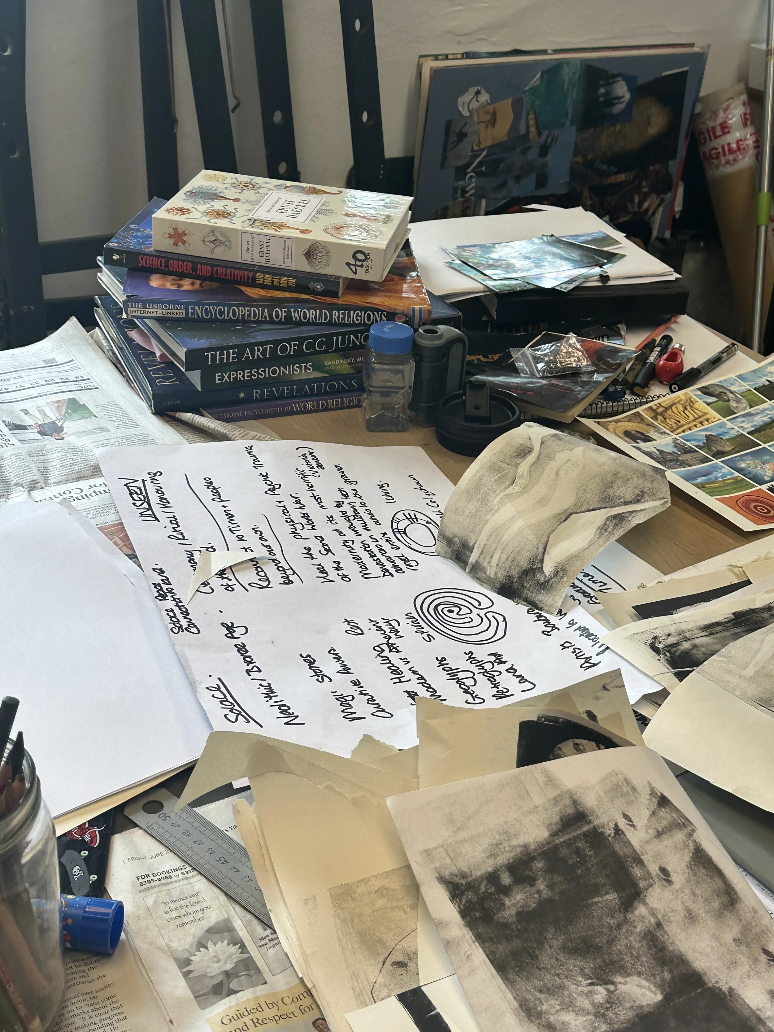 A desk full of ideas,  books, sketches, papers, photographs, and art supplies scattered across the surface in a workspace.