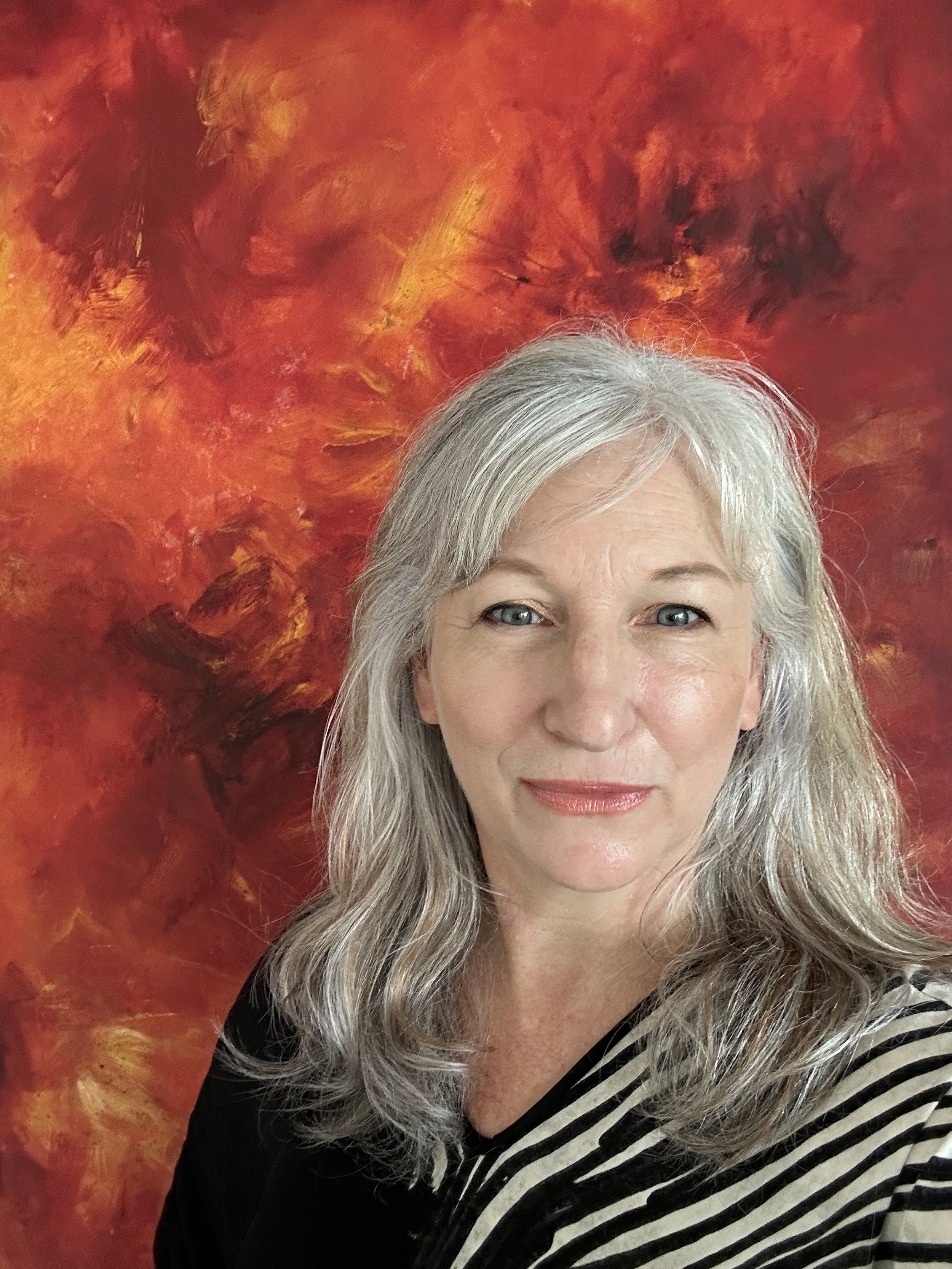 A woman with gray hair and blue eyes smiling, wearing a black and beige striped top, standing in front of an abstract fiery orange and red background.