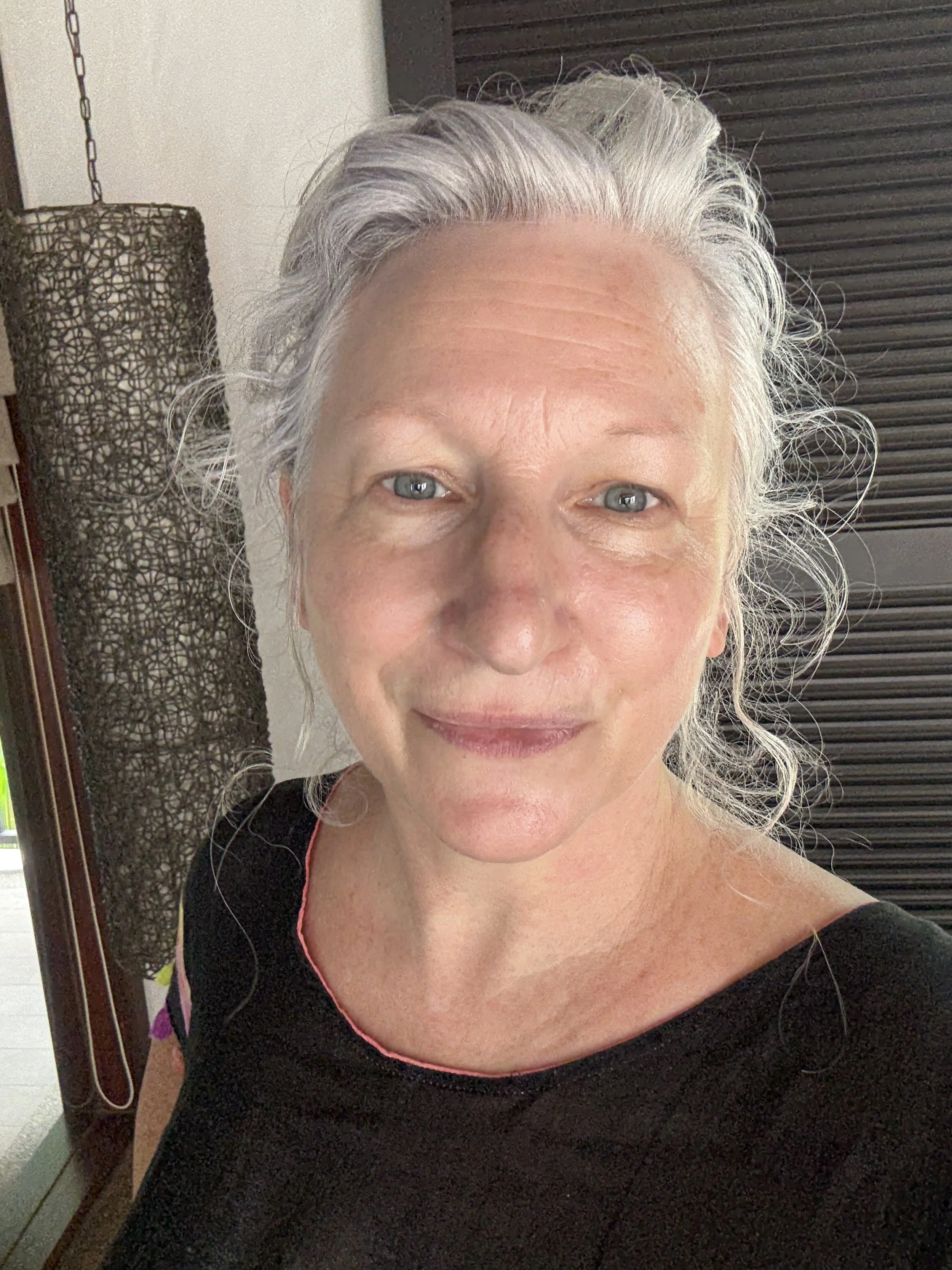 Hilary Lee smiling at the camera, wearing a black top, indoors with a decorative wall lamp and dark shutters in the background.