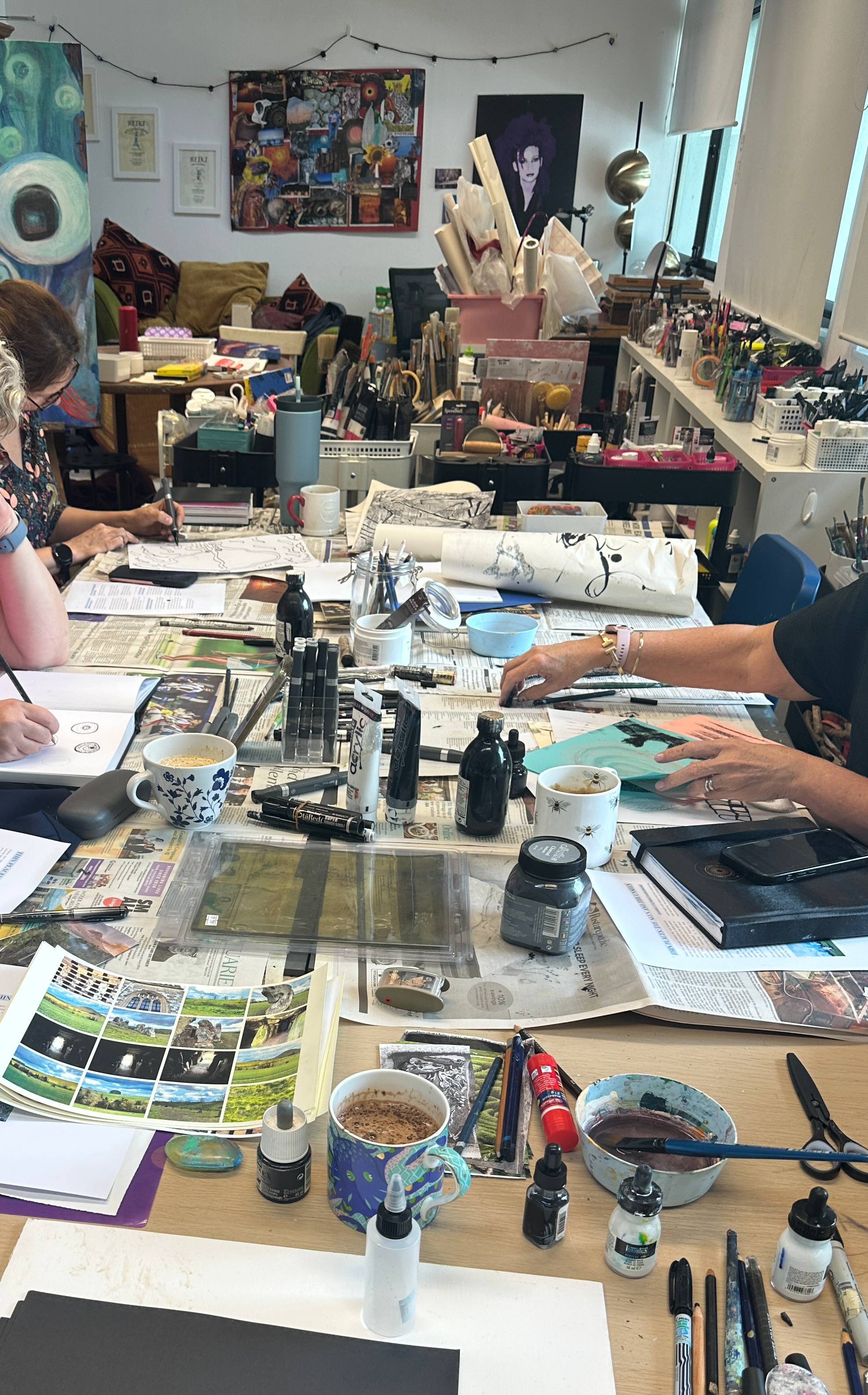 Art studio workspace with painting supplies, notebooks, brushes and paper  and two women working on art projects. The chaotic background features artwork, creativity, and windows letting in natural light.