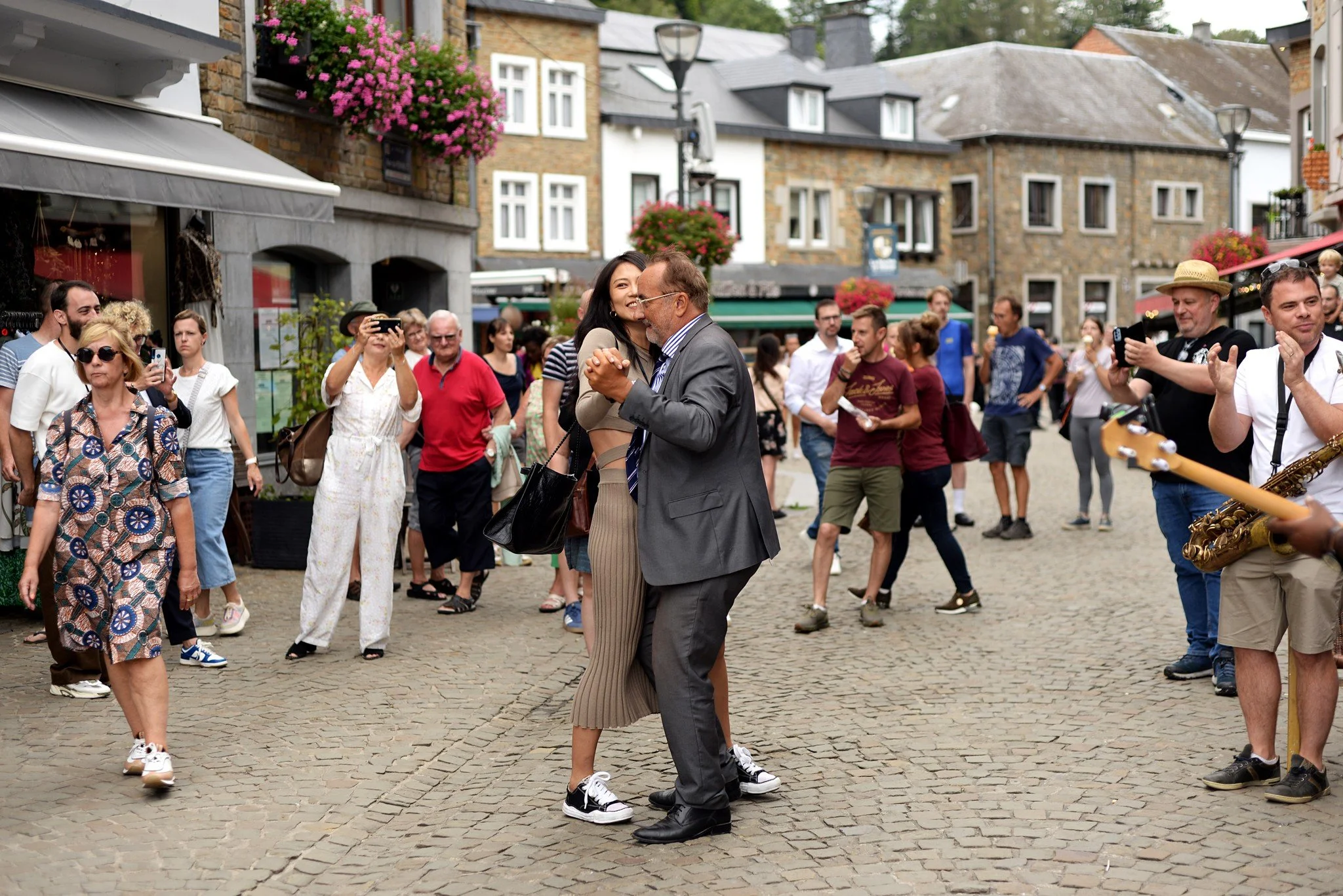 Deux personnes dansent au centre d'une place pavée entourée de nombreux spectateurs. La scène évoque une ambiance festive avec un groupe de musiciens à droite, certains prenant des photos ou vidéos. L'arrière-plan montre des bâtiments élégants avec d