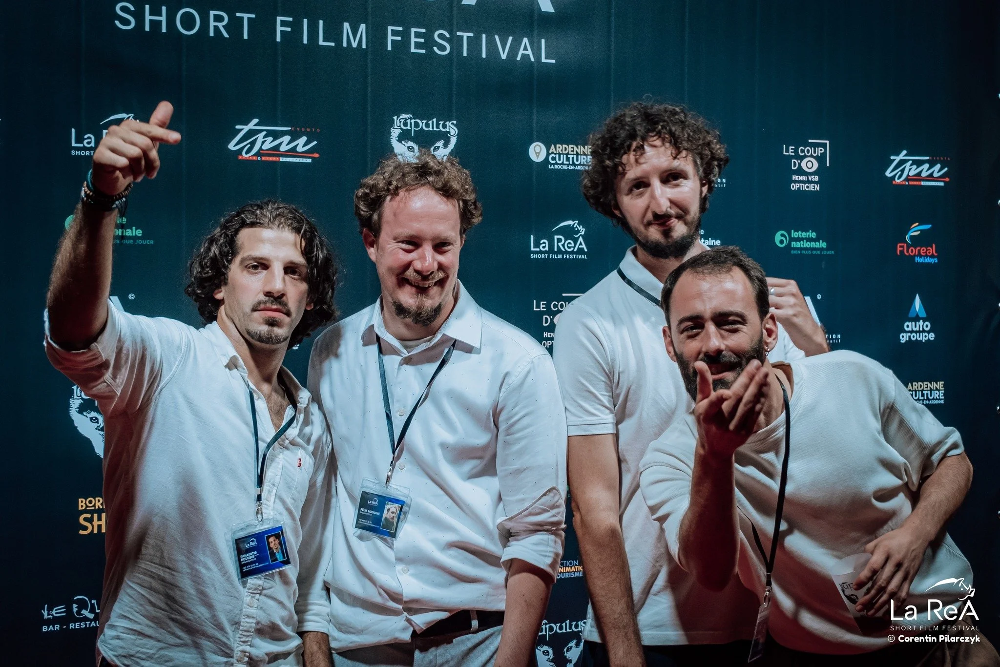 Quatre hommes posent devant un fond noir avec des logos pour un festival de courts métrages, certains sourient et font des gestes avec leurs mains.