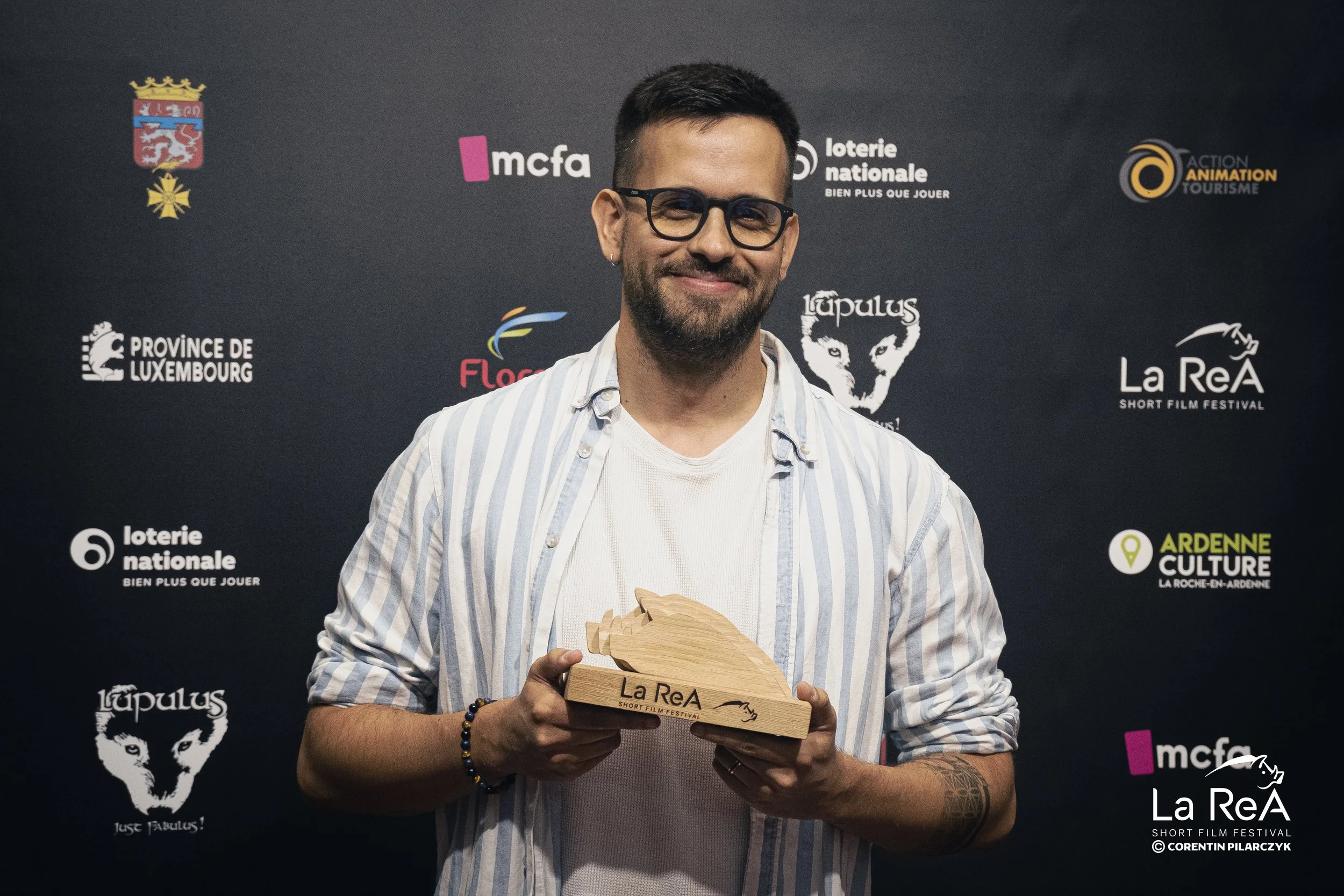 Homme souriant tenant un trophée en forme de tête de loup en bois lors du festival du court métrage La ReA, avec derrière lui un mur de logos d'organisations et d'événements.