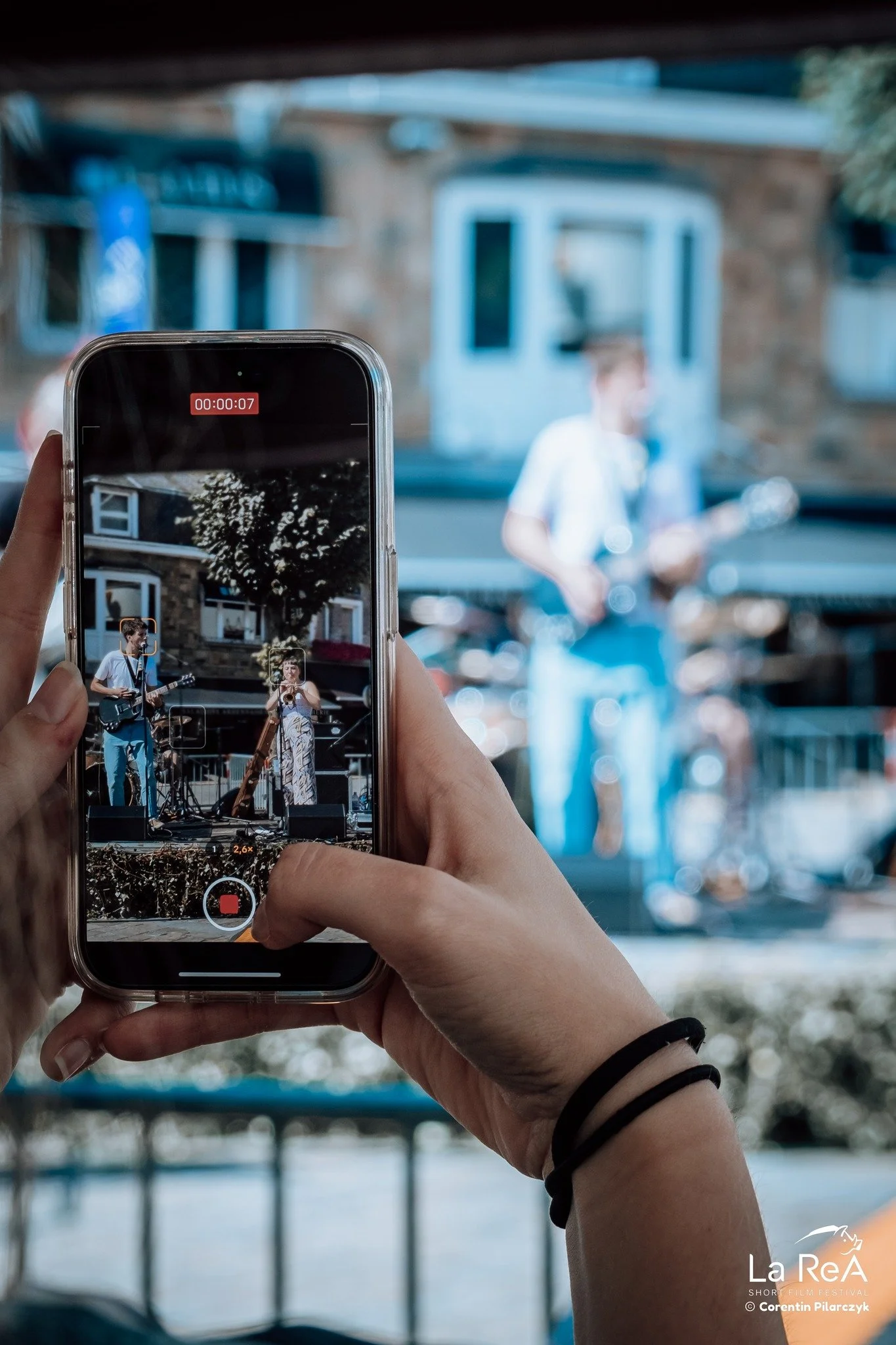 Une personne en train d'enregistrer un groupe de musique jouant en extérieur lors d'un festival. La scène est vue à travers l'écran d'un téléphone portable.