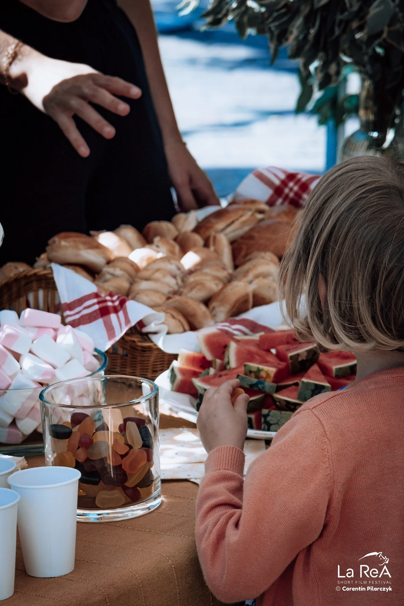 Un enfant mangeant des tranches de pastèque à une table avec des bonbons, marshmallows et pâtisseries, lors d'un festival en plein air.