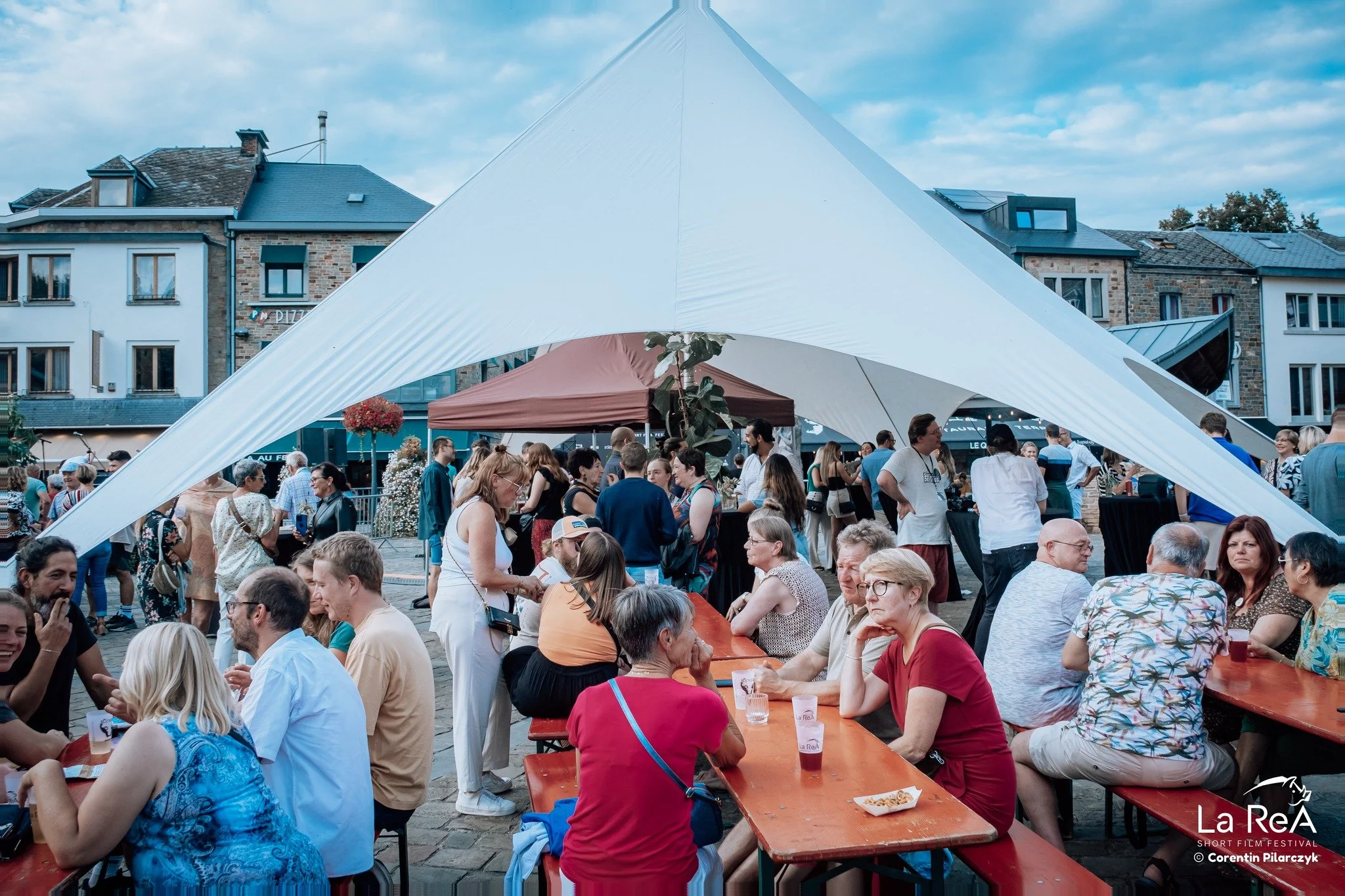 Une foule de personnes assises à des tables en bois dehors lors d’un festival ou d’une fête, sous une grande tente blanche. En arrière-plan, des bâtiments en pierre et en béton, et un ciel partiellement nuageux.