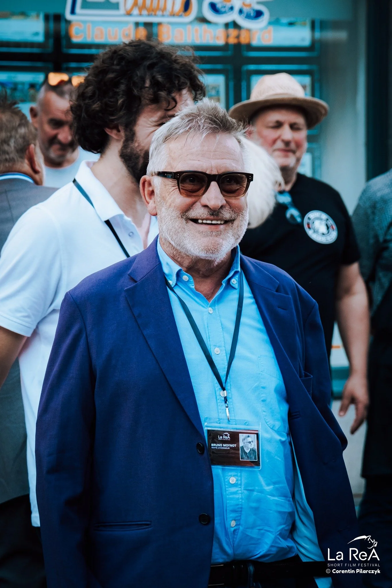 Groupe de personnes souriantes lors d'un événement, l'homme au centre porte une veste bleue et des lunettes de soleil, portant un badge avec le nom Bruno Moynot, au festival de courts-métrages La Rea.
