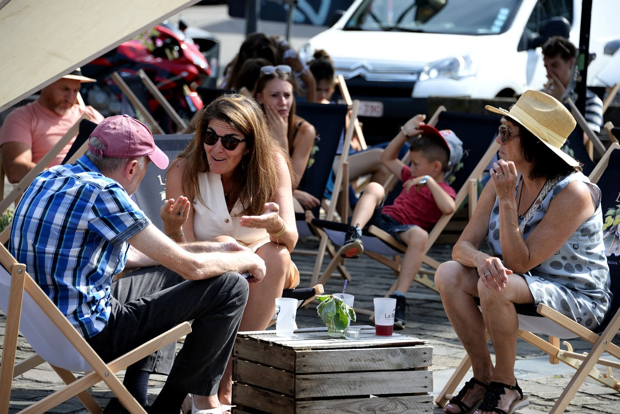 Groupe de personnes assises en plein air, discutant et profitant d'une journée ensoleillée, avec des voitures en arrière-plan.