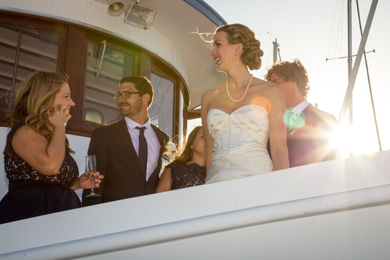 A wedding celebration on a yacht with smiling guests, including a bride in a white dress and a woman in a dark dress holding a champagne glass, with sunlight shining in the background.