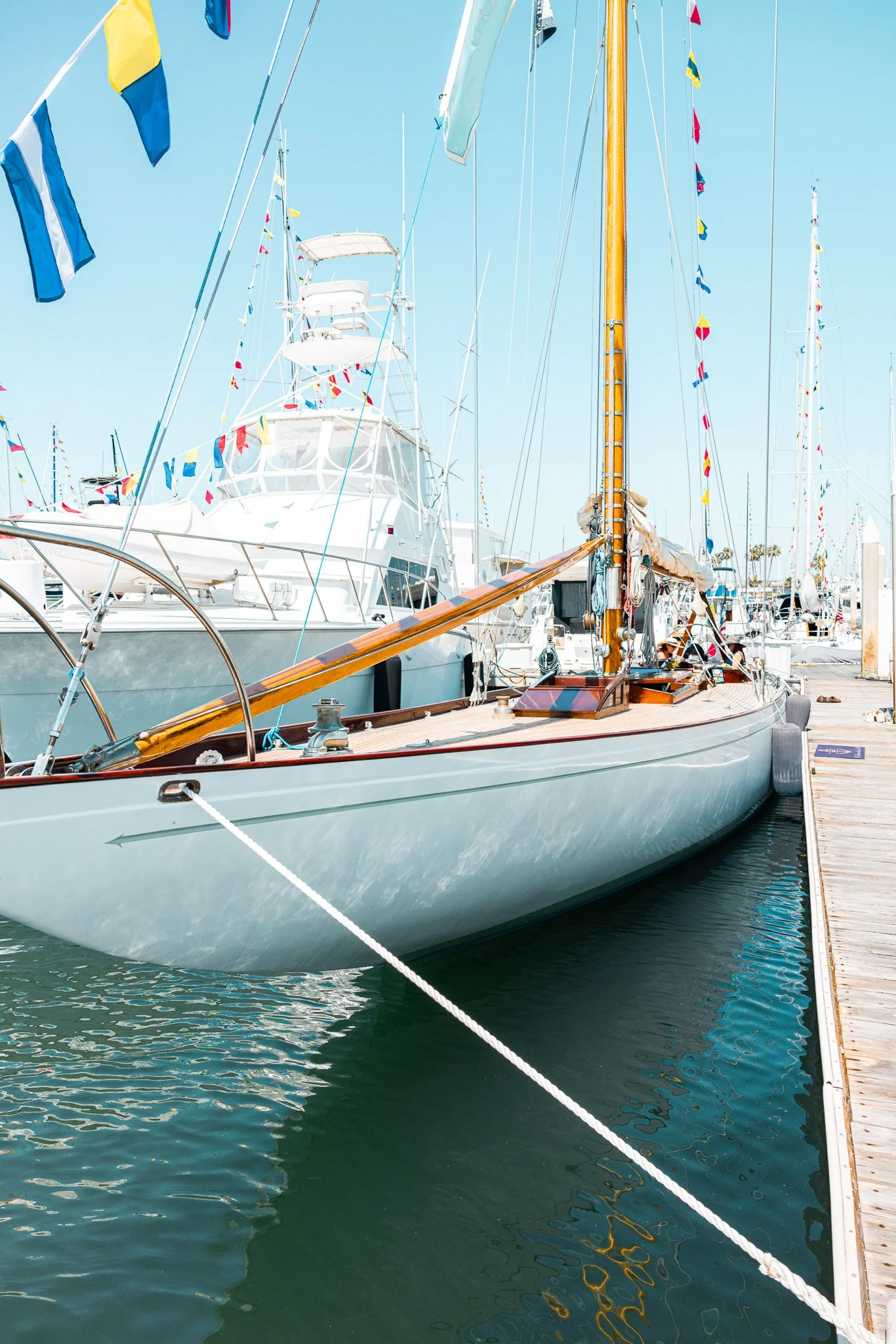 A sailboat moored at a marina with multiple other boats, colorful flags hanging from the masts, and clear blue skies.