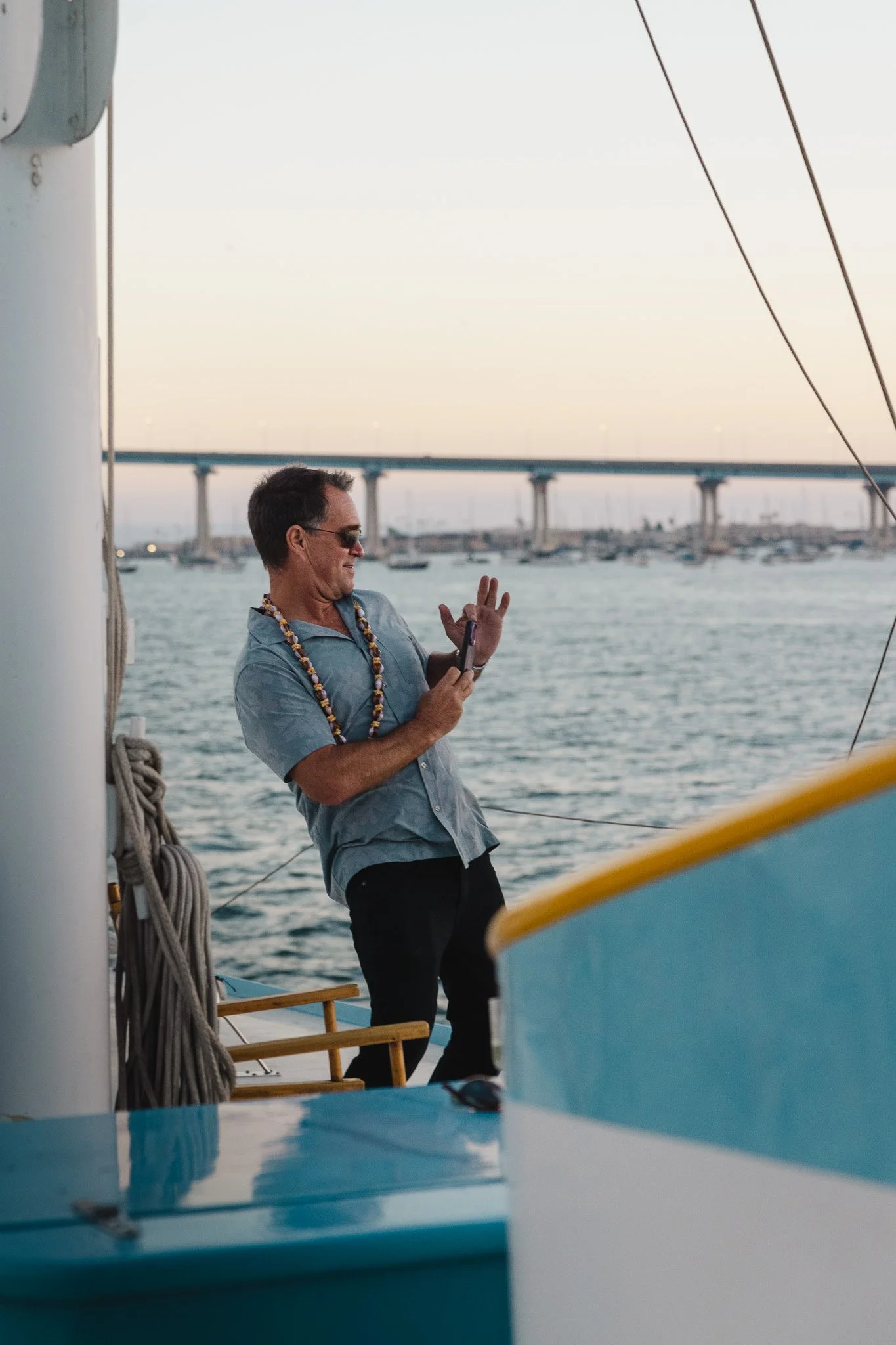 Man on a boat taking a selfie with water and boats in the background at sunset.