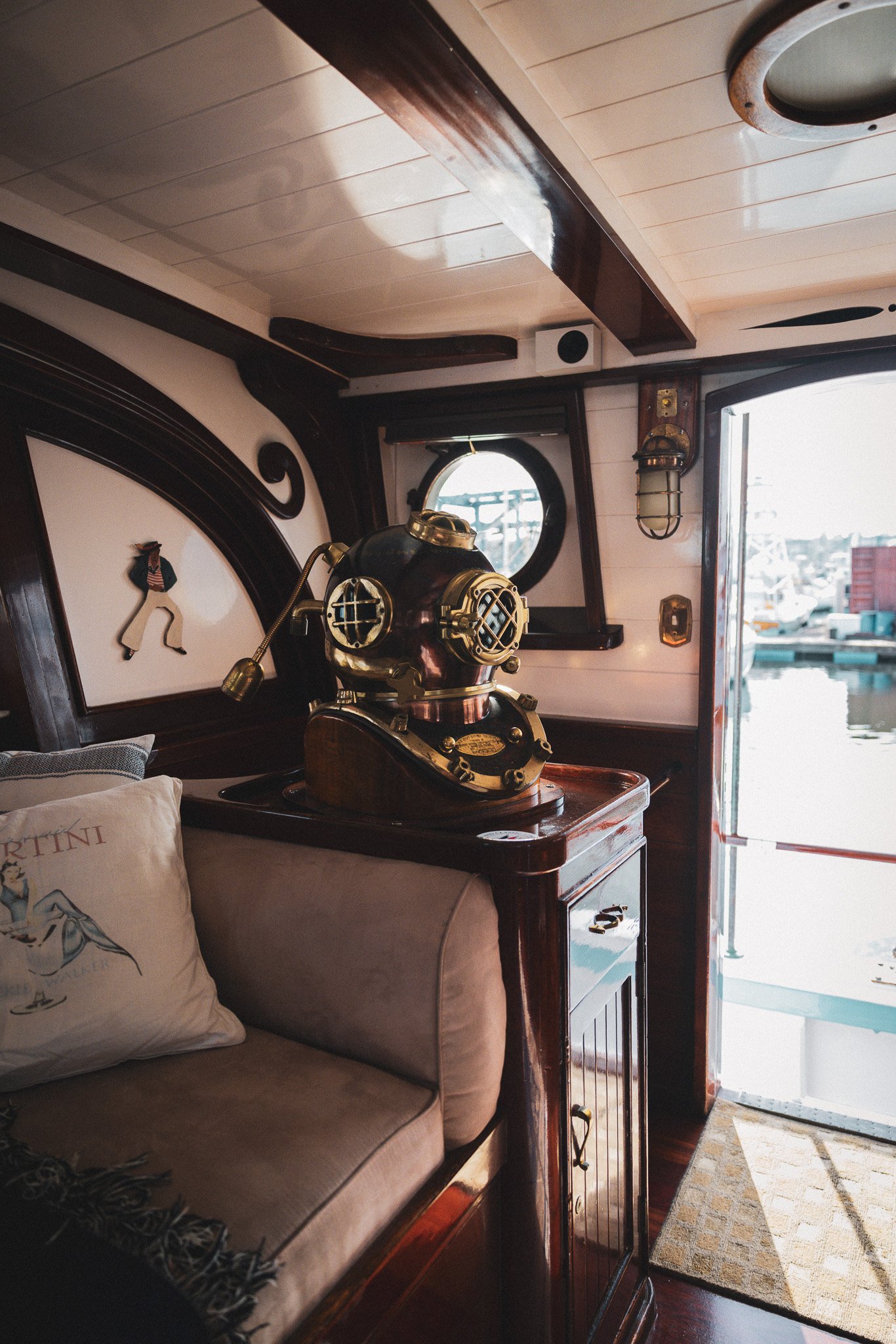 Interior of a boat with a vintage bronze diving helmet on a wooden cabinet, window with water view, and nautical decor.