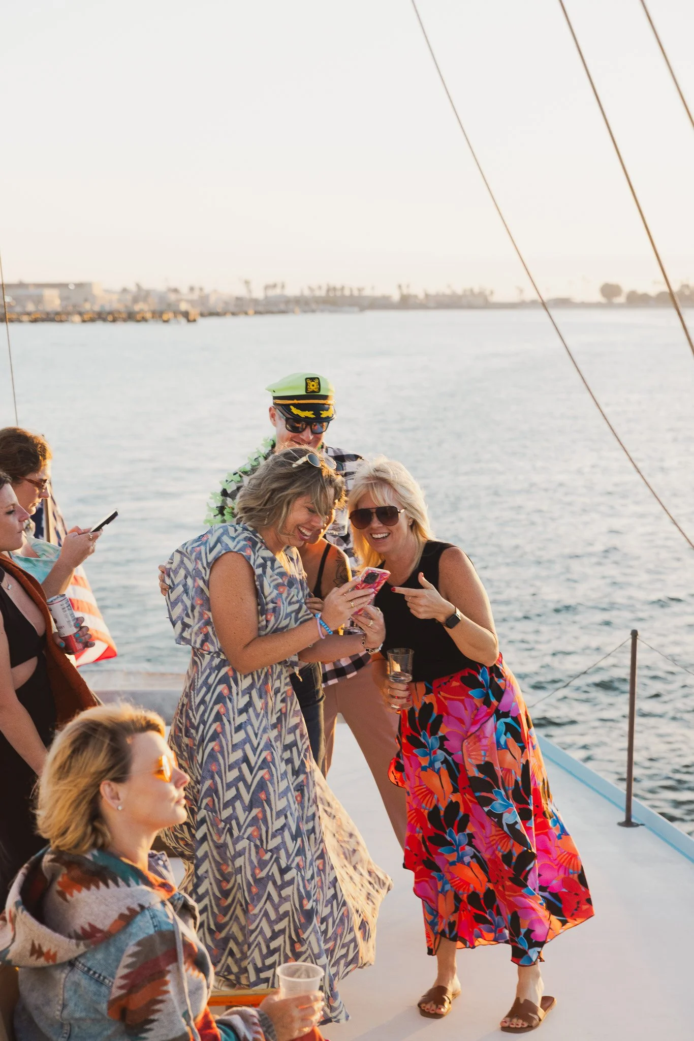 Group of people enjoying a boat party by the water at sunset, some taking photos and holding drinks, wearing summer clothing and sunglasses.