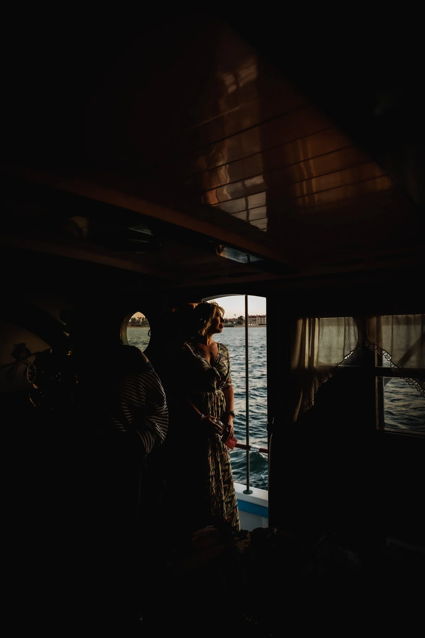 A woman looks out from the window of a boat at sunset, with water visible outside and interior dimly lit.