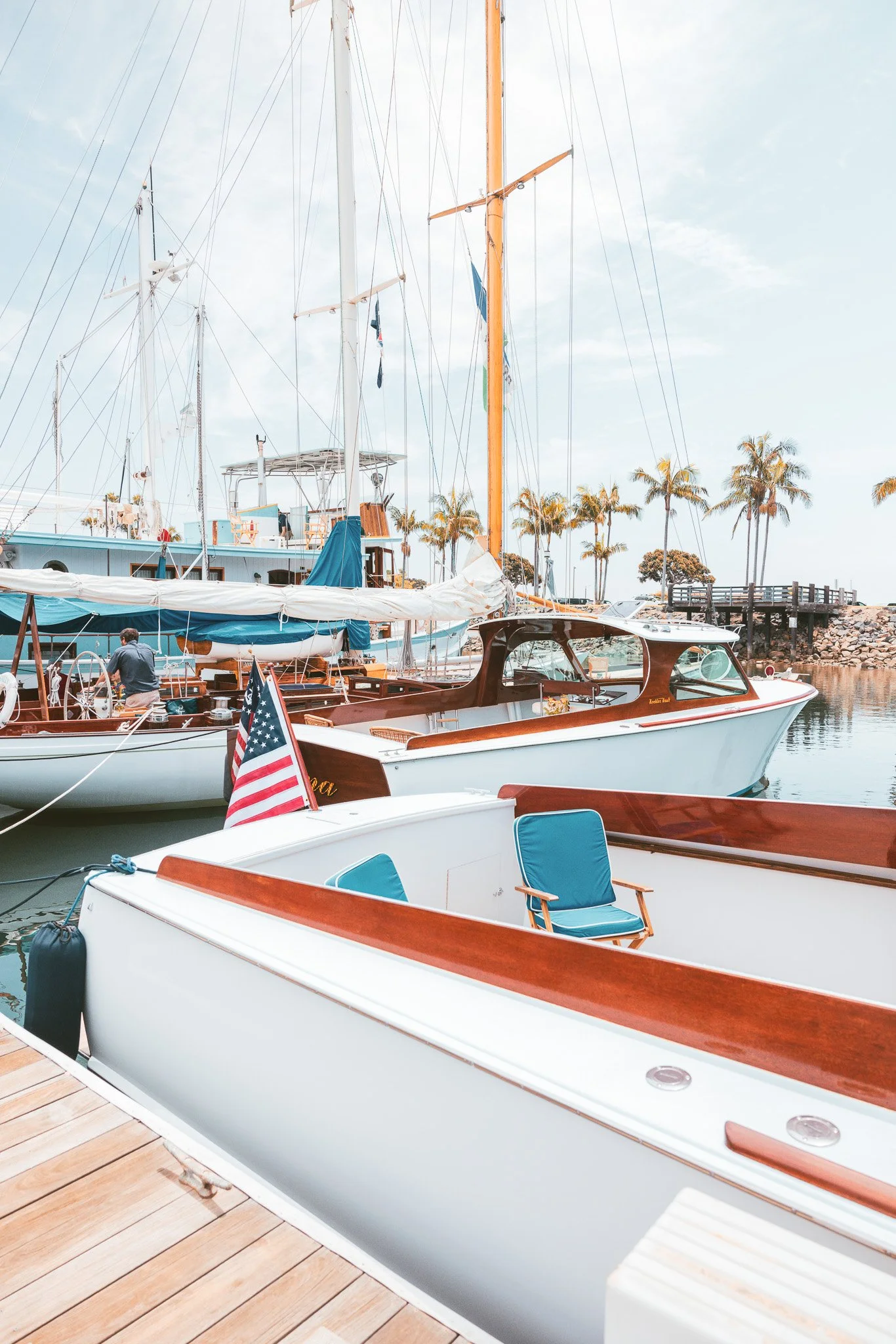 Boats docked at a marina with palm trees and a walkway in the background on a partly cloudy day.
