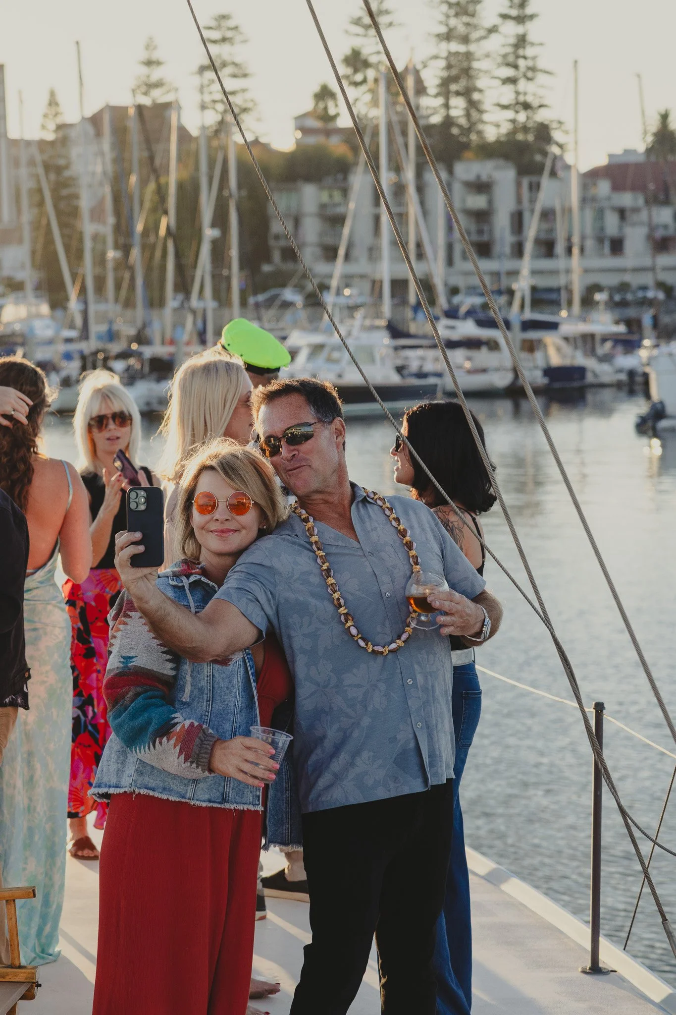 People on a boat taking a selfie with sailboats and a marina in the background during sunset.