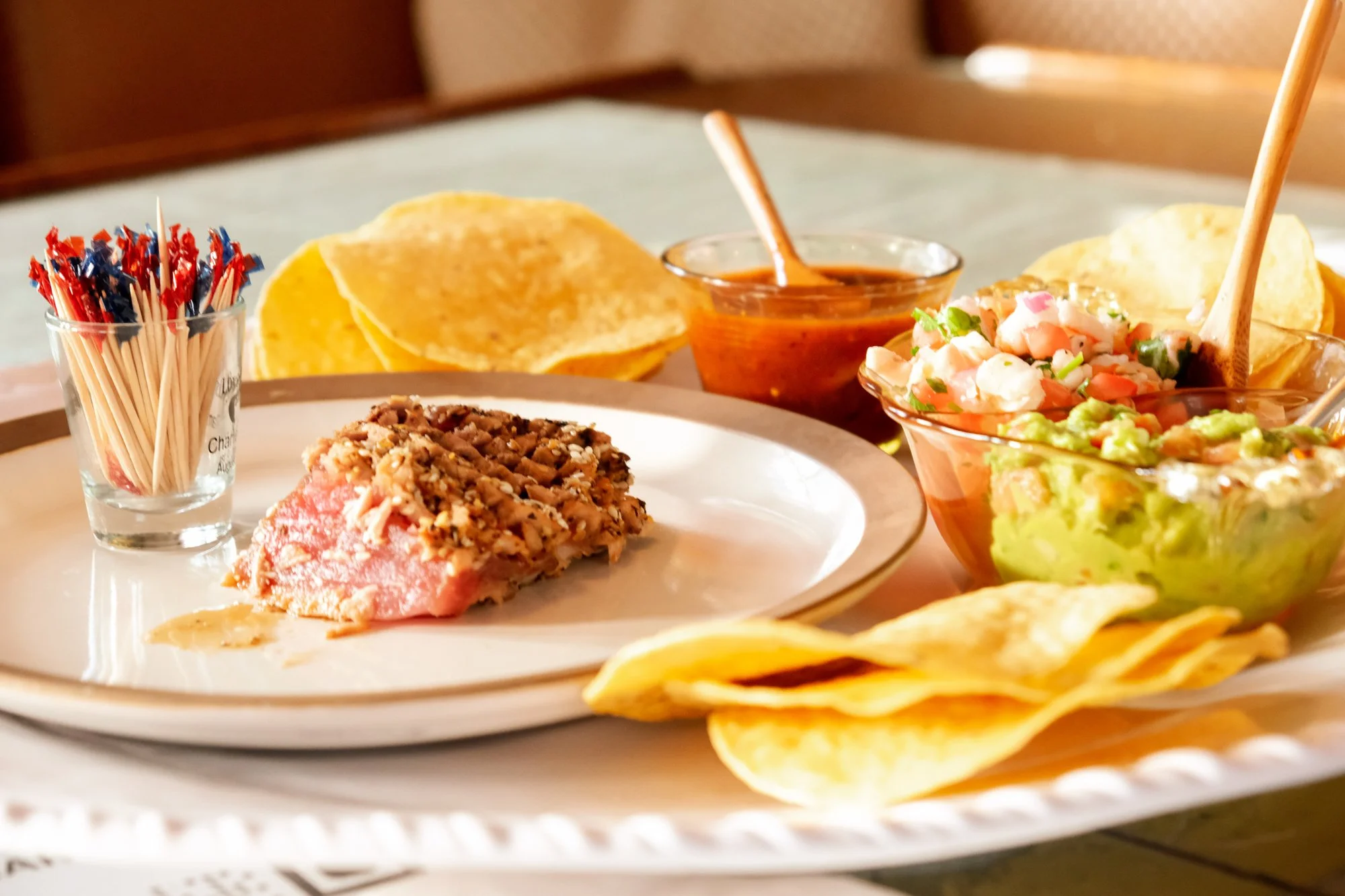 Plate with a raw piece of meat topped with chopped nuts, tortilla chips, small bowls of salsa, guacamole, and pico de gallo, and a glass of toothpicks.