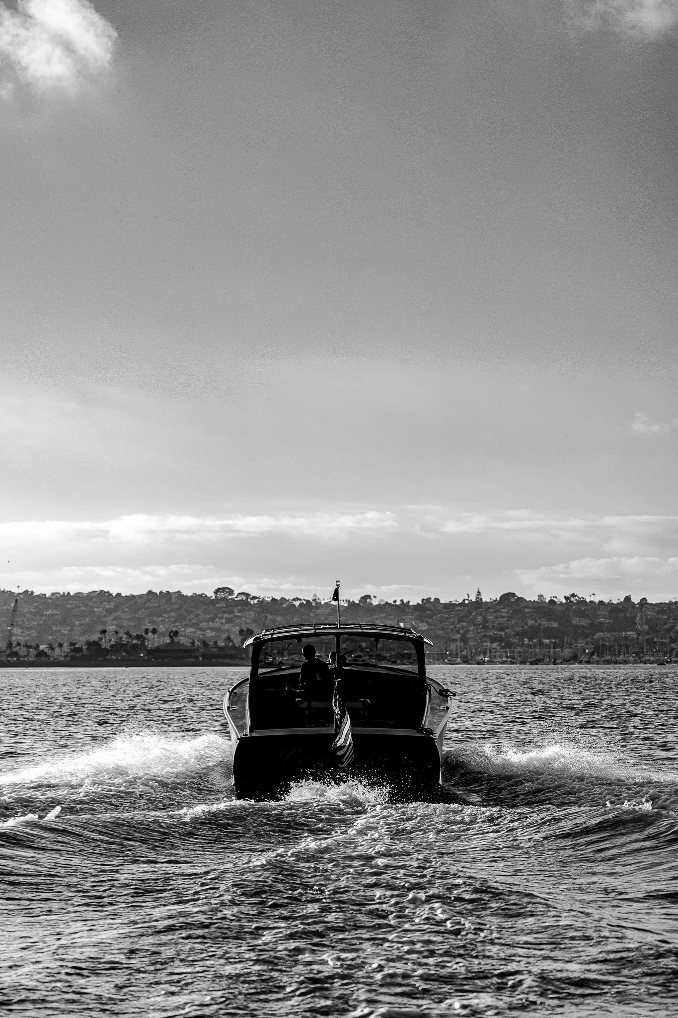 A boat moving away from the camera on the water with a person on board. The background features a distant shoreline with trees and buildings, under a mostly cloudy sky.