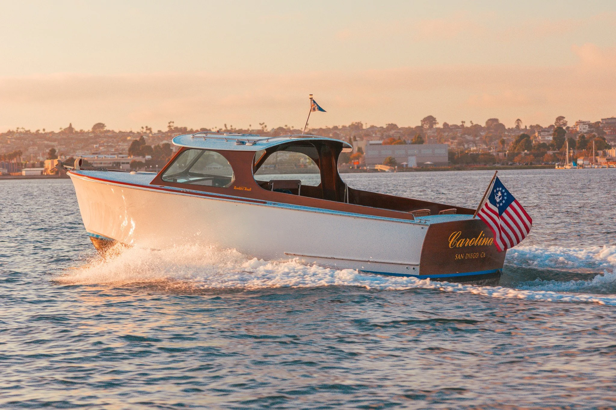 A white motorboat with a brown roof moving across the water near San Diego, California, at sunset, with an American flag at the back of the boat.
