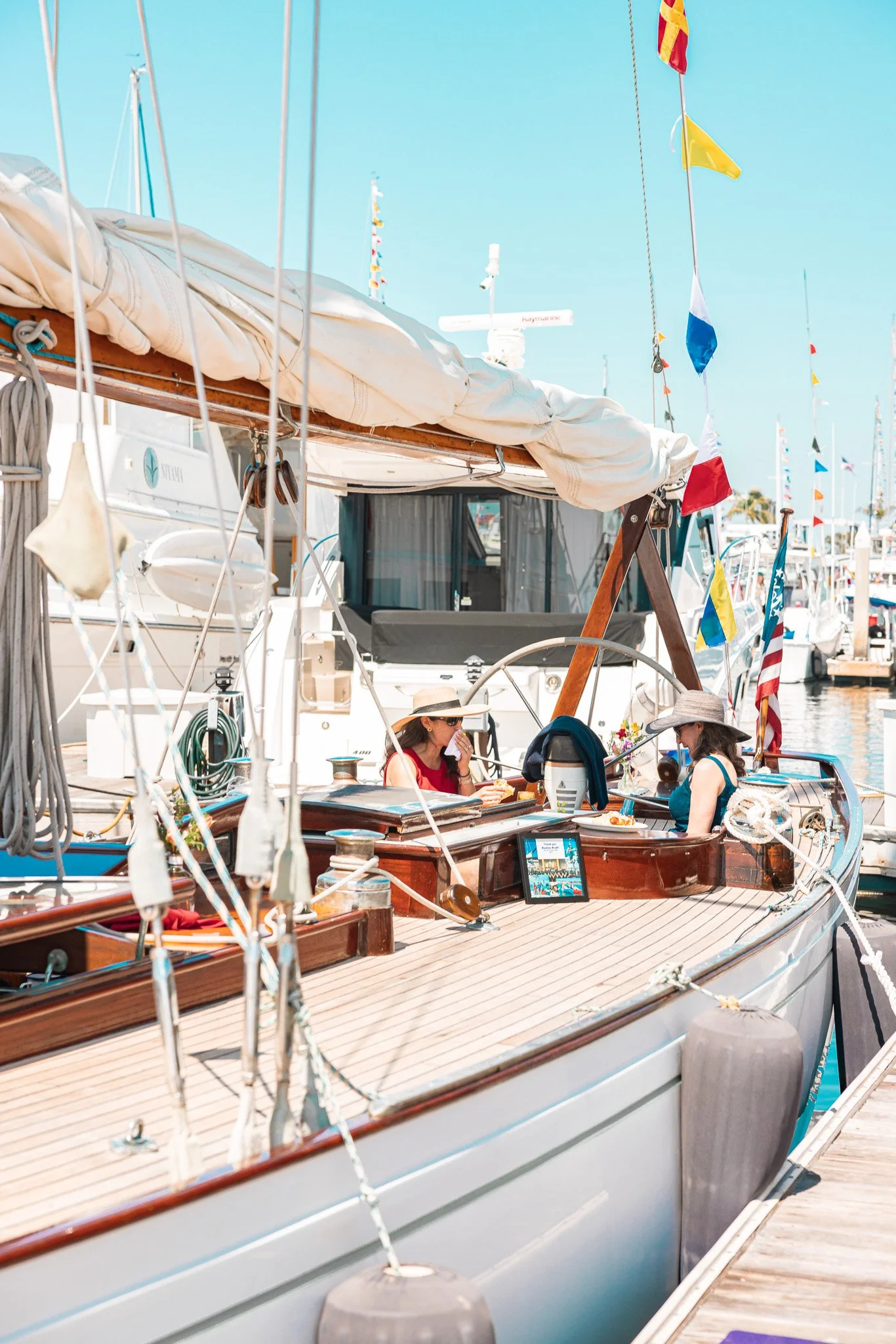 Two women sitting on a boat at a marina, wearing hats, enjoying drinks and snacks, with various boats and colorful flags in the background.