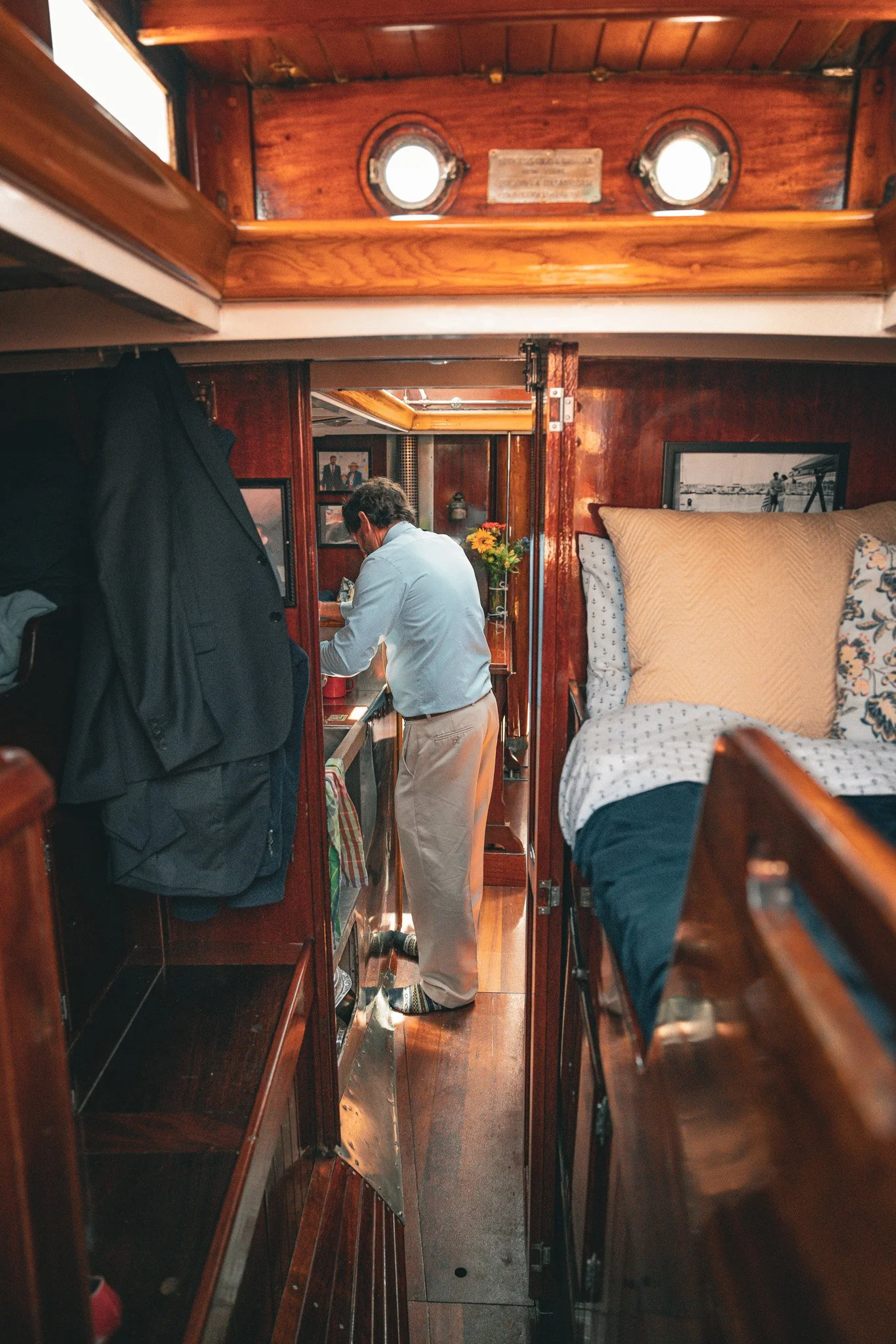 Interior of a boat cabin with a man in a light blue shirt at a small kitchen sink area, and a bed with pillows to the right.