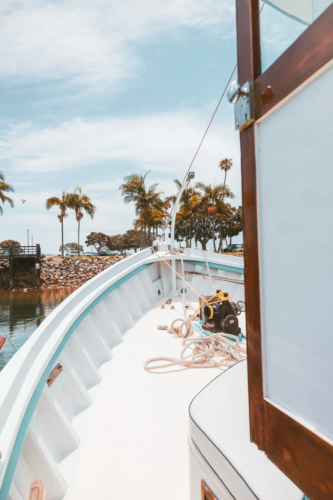 View of a boat's bow with ropes and equipment, next to a wooden structure, with palm trees and a clear sky in the background.