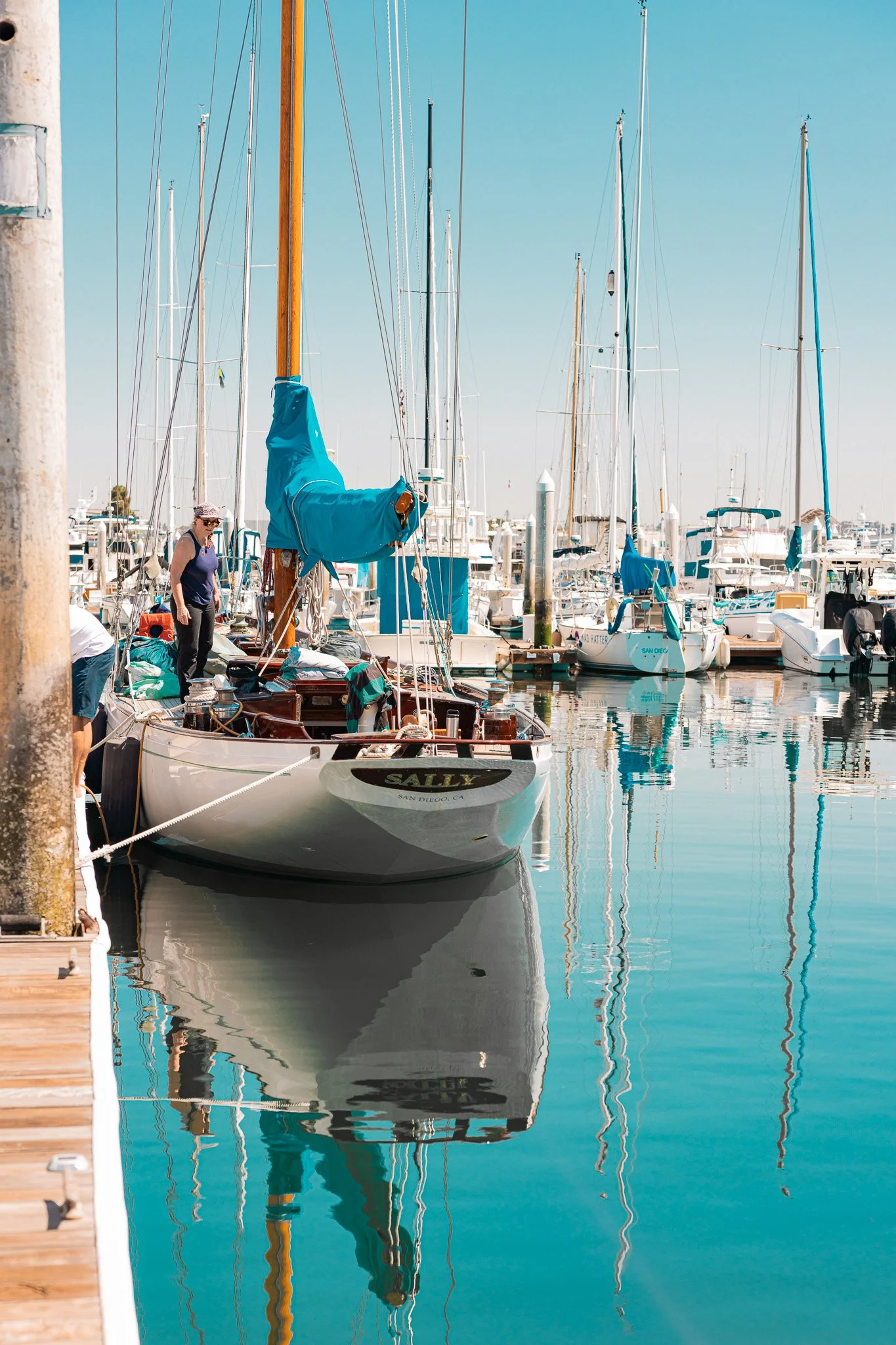 A marina filled with sailboats docked along the pier, with one sailboat named Sally in the foreground and others in the background, reflected in calm water on a sunny day.