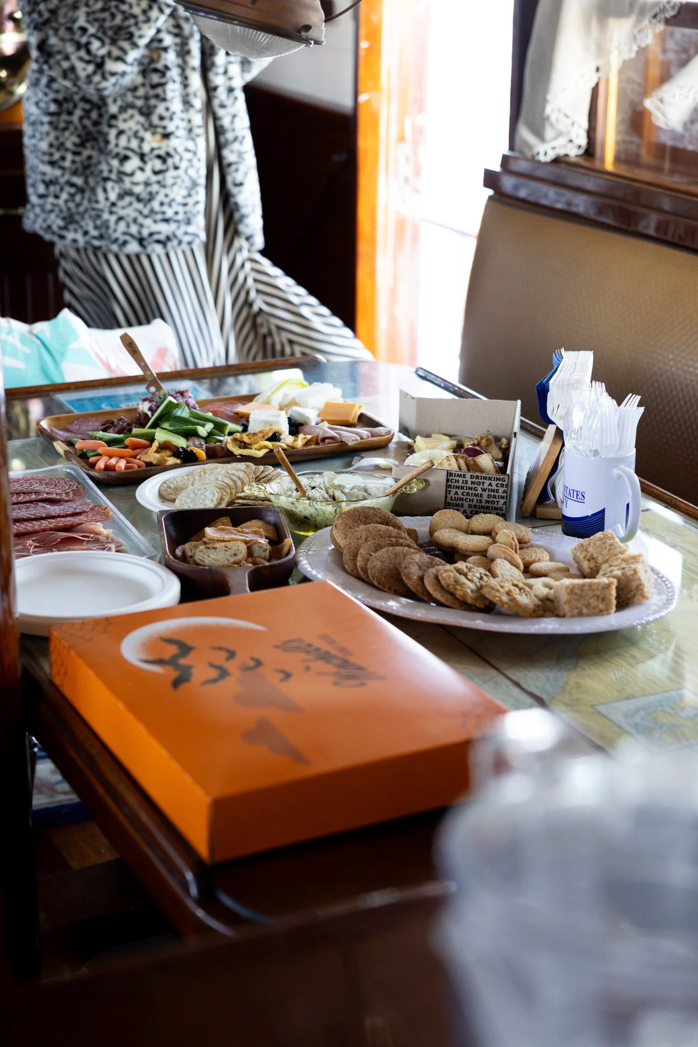 A table set for a breakfast or brunch with various foods including cold cuts, cheeses, crackers, cookies, vegetables, and a box with a Halloween theme. There are plastic utensils in a cup, and a person in a patterned outfit standing nearby, with a wi